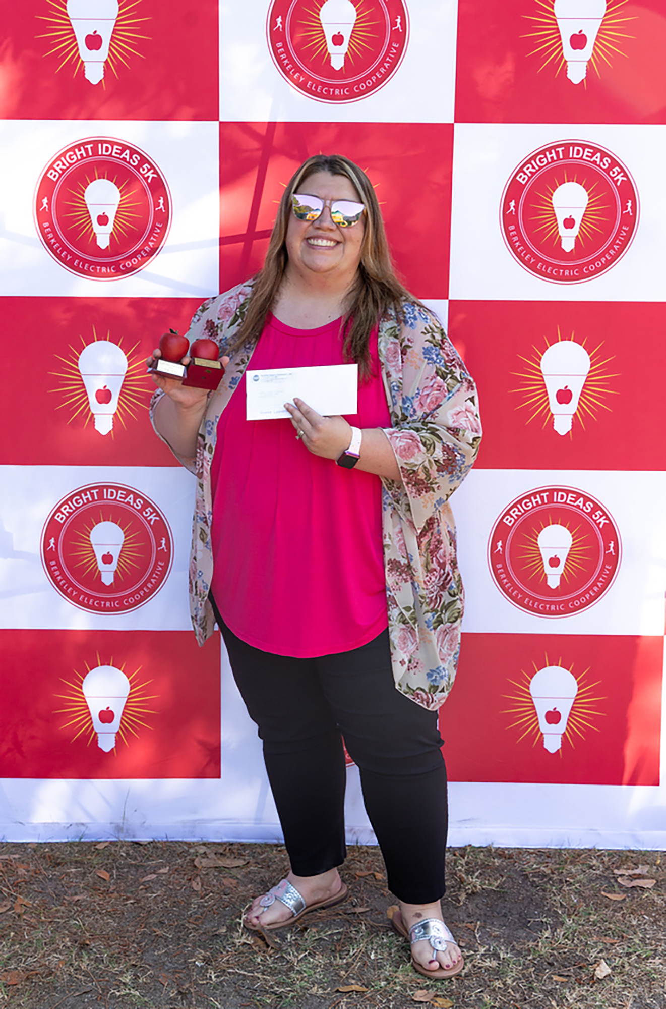 Woman in a pink shirt holds a certificate and a trophy, standing before a red and white wall with light bulb logos.