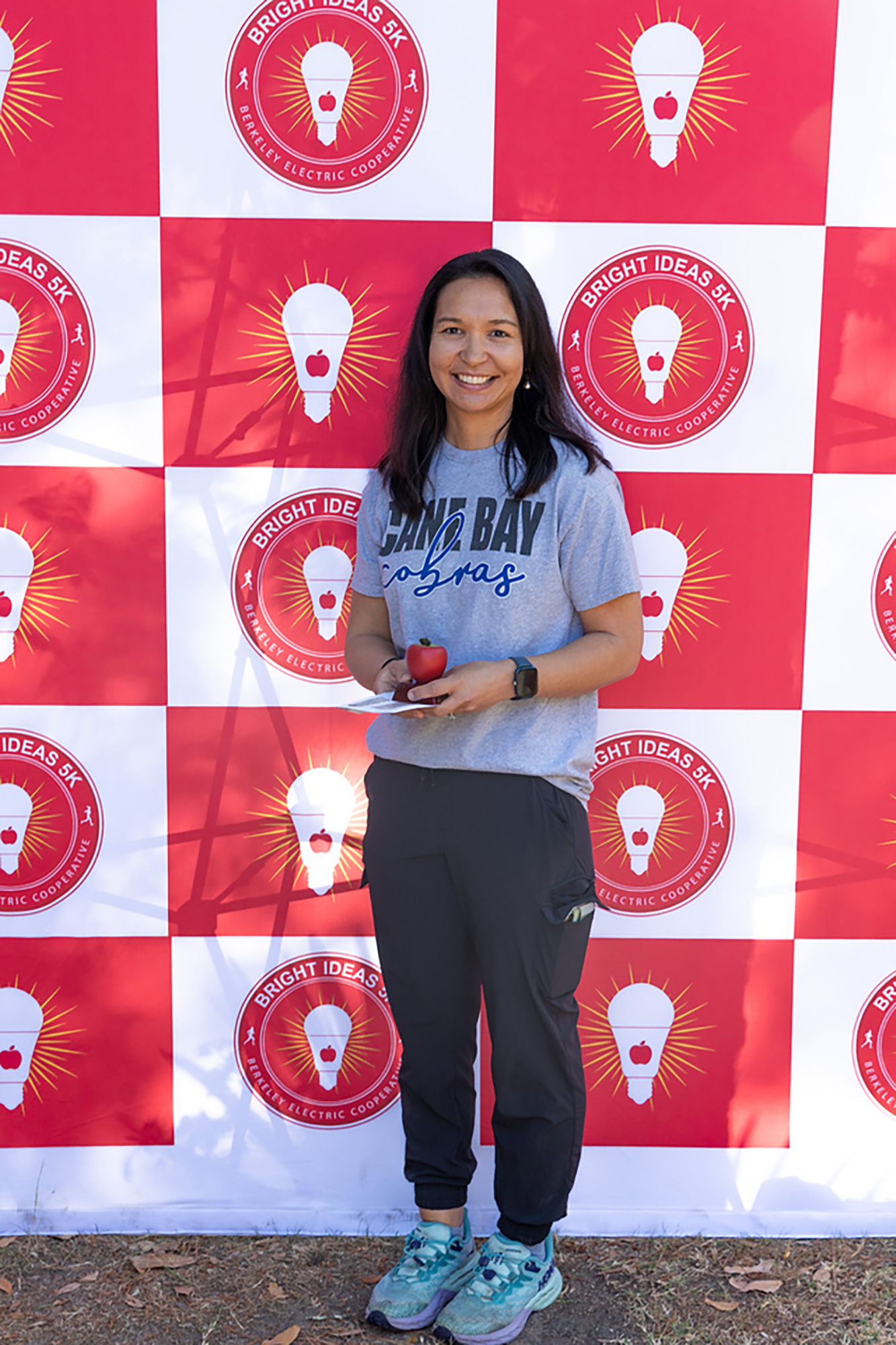 Person in athletic clothes holds a red apple in front of a red and white checkered wall with light bulb logos.