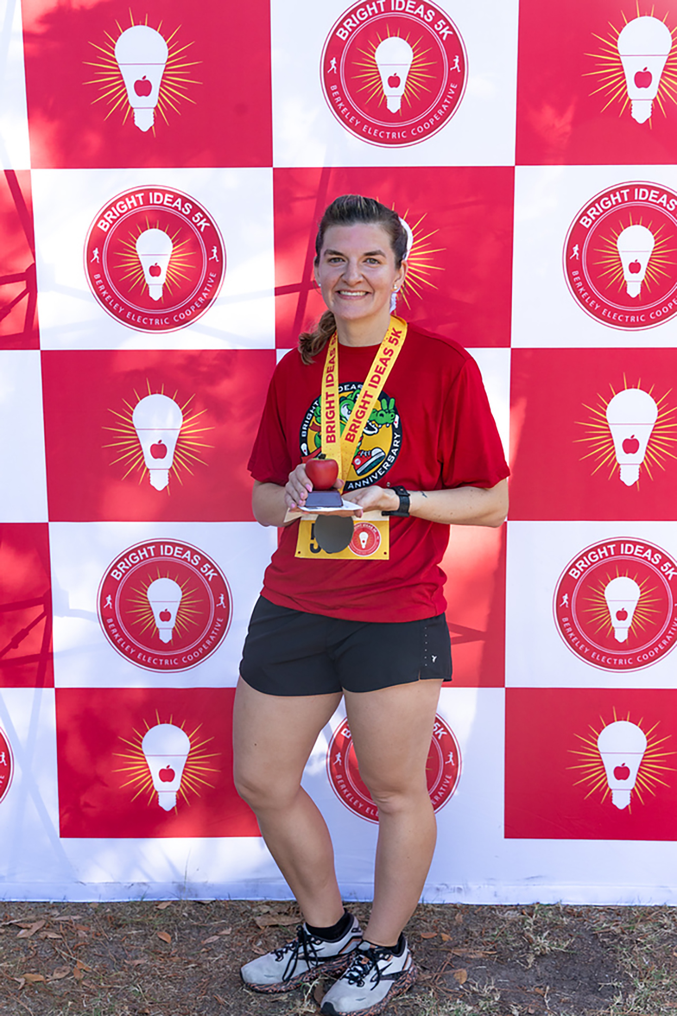 Person in red shirt holding medal standing in front of red and white checkered wall with light bulb logo.