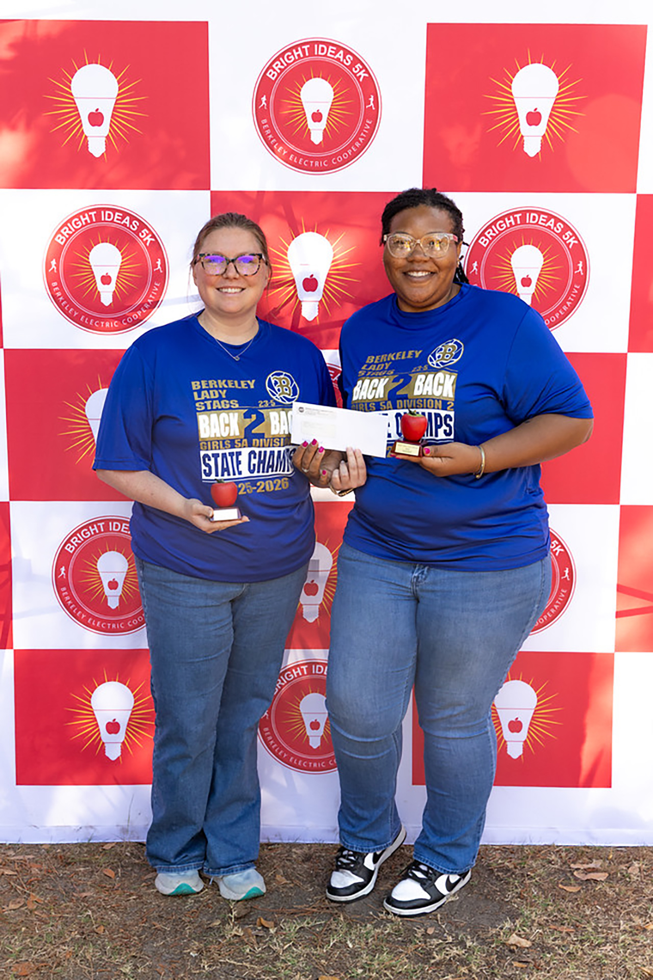 Two women in blue shirts hold awards and apples in front of a red and white wall with light bulb logos.