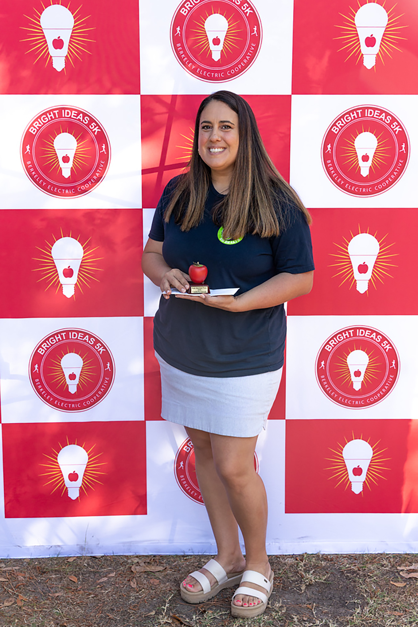 A woman stands before a wall of red and white squares, holding a plate with a red apple.