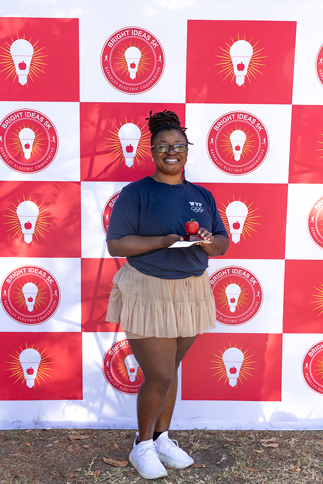 A woman stands against a red-and-white wall with light bulb logos, holding a small apple.