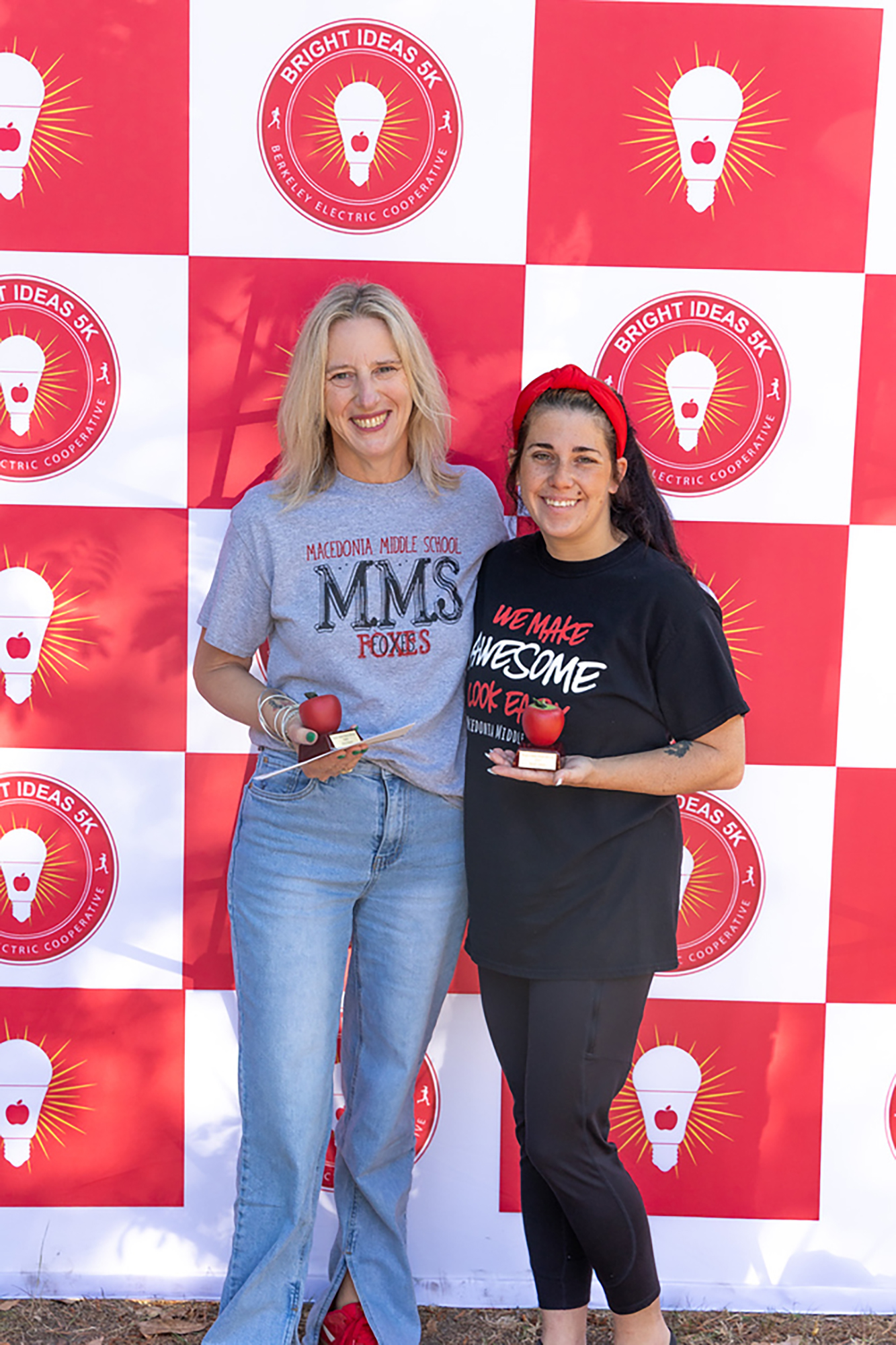 Two women smile before a backdrop with red and white squares and lit lightbulb logos. One holds an apple.