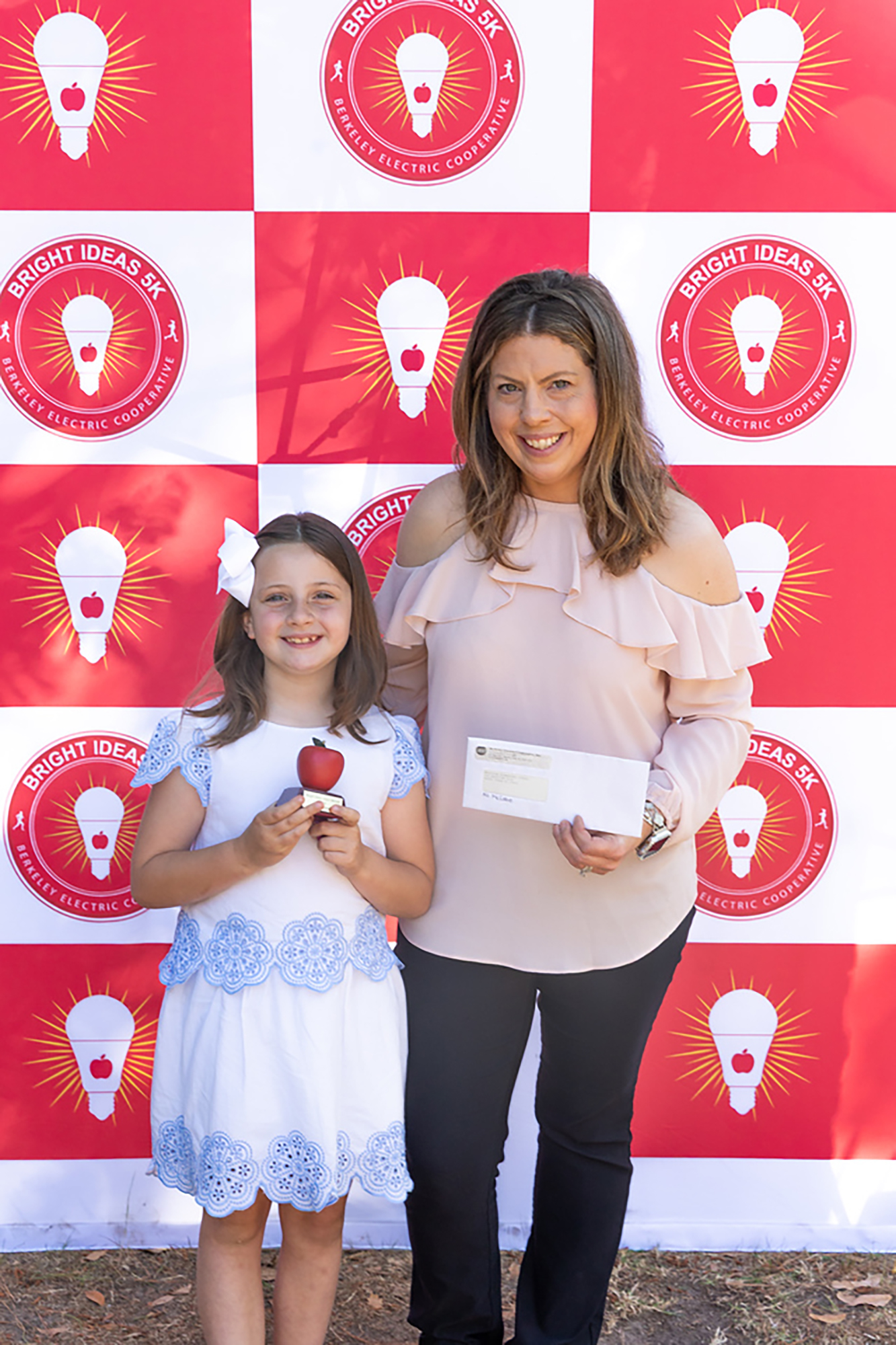 A woman and a girl stand together in front of a red and white wall with light bulbs.