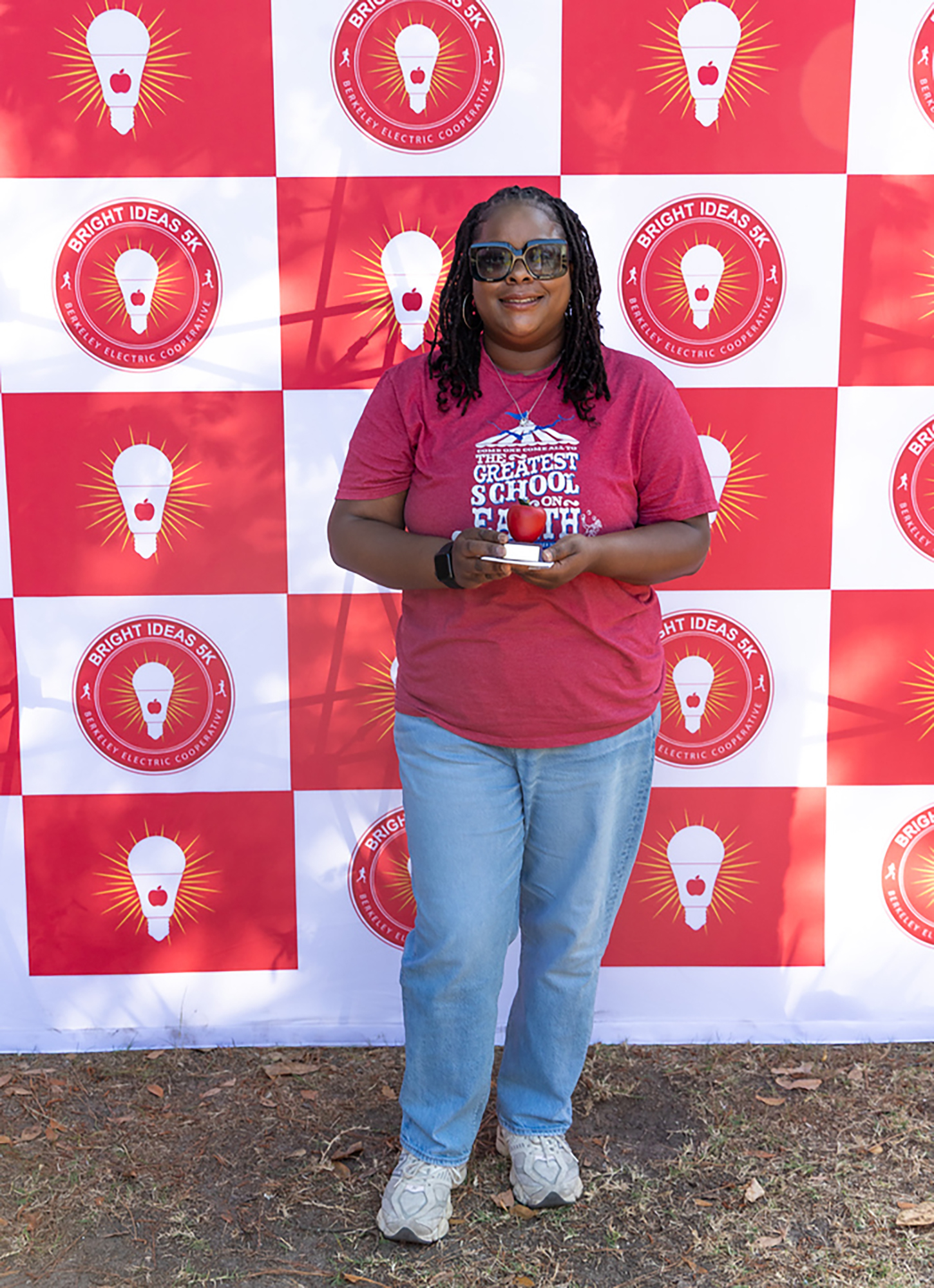 Person stands before a wall with red and white squares, each containing a light bulb. Person holds an award and wears sunglasses, a maroon shirt, blue jeans, and white shoes.