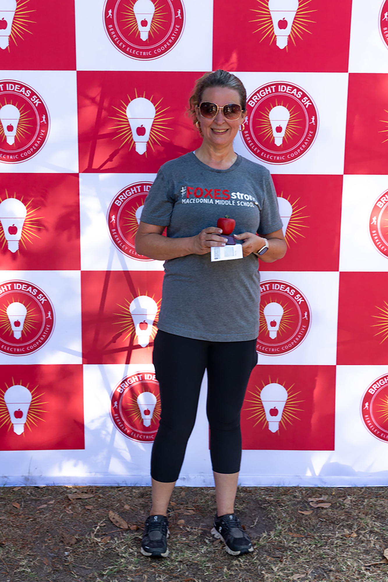 A woman stands in front of a red and white background with light bulbs, wearing a gray shirt and black leggings. She holds a small object and smiles.
