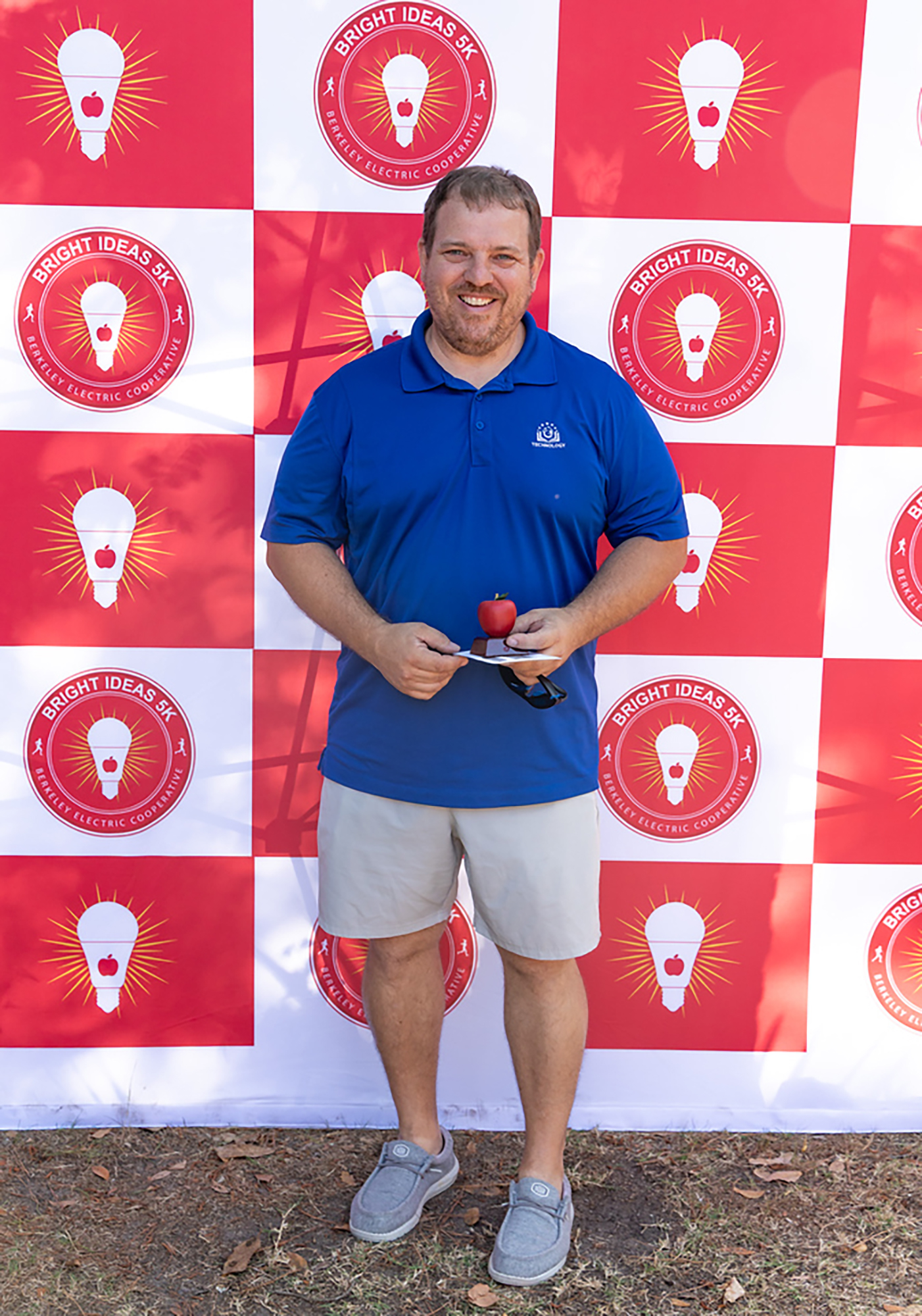 Person wearing a blue shirt and khaki shorts holding a red apple in front of a red and white patterned backdrop.