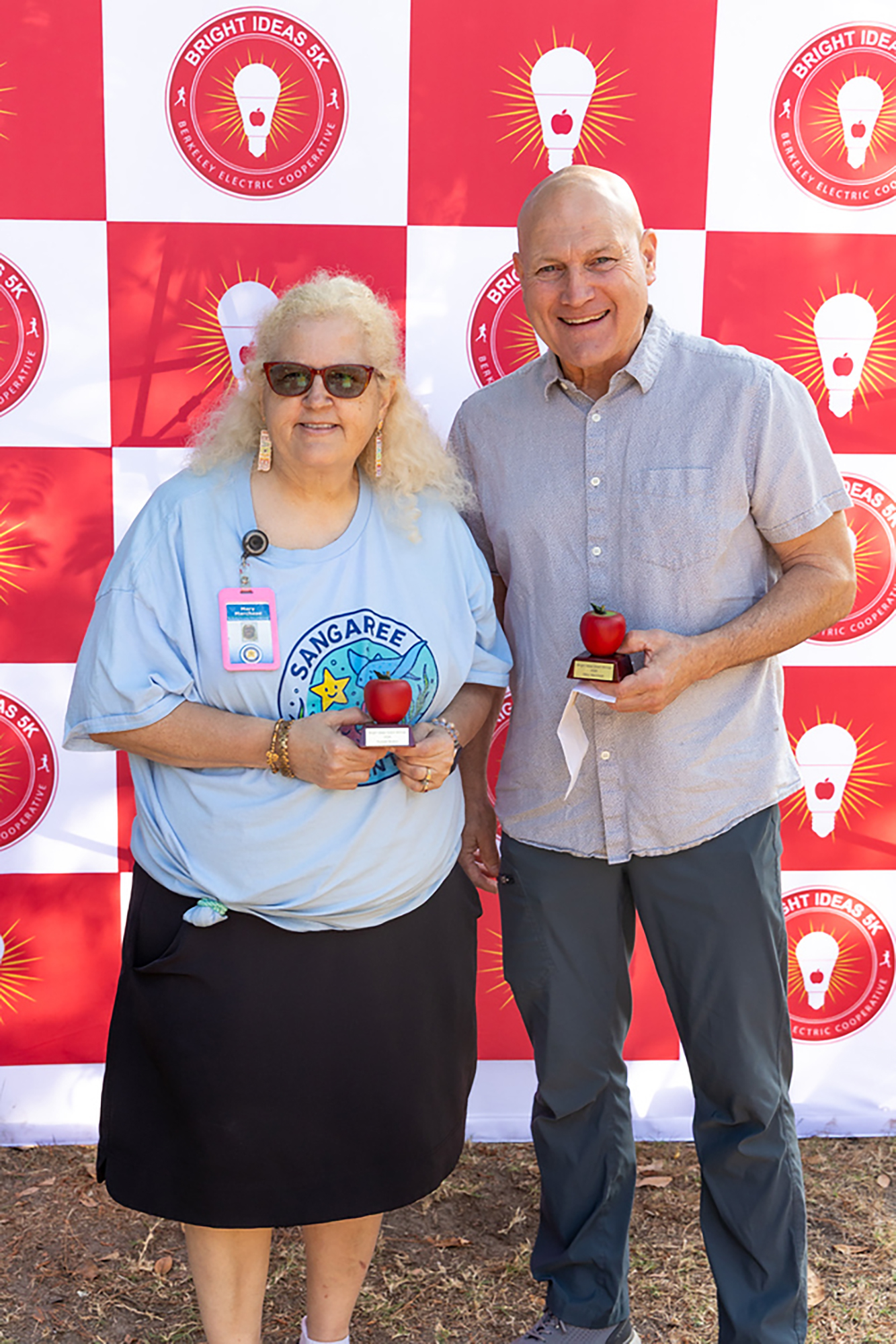 Two people stand in front of a red and white backdrop. Both hold awards, and the woman wears sunglasses.