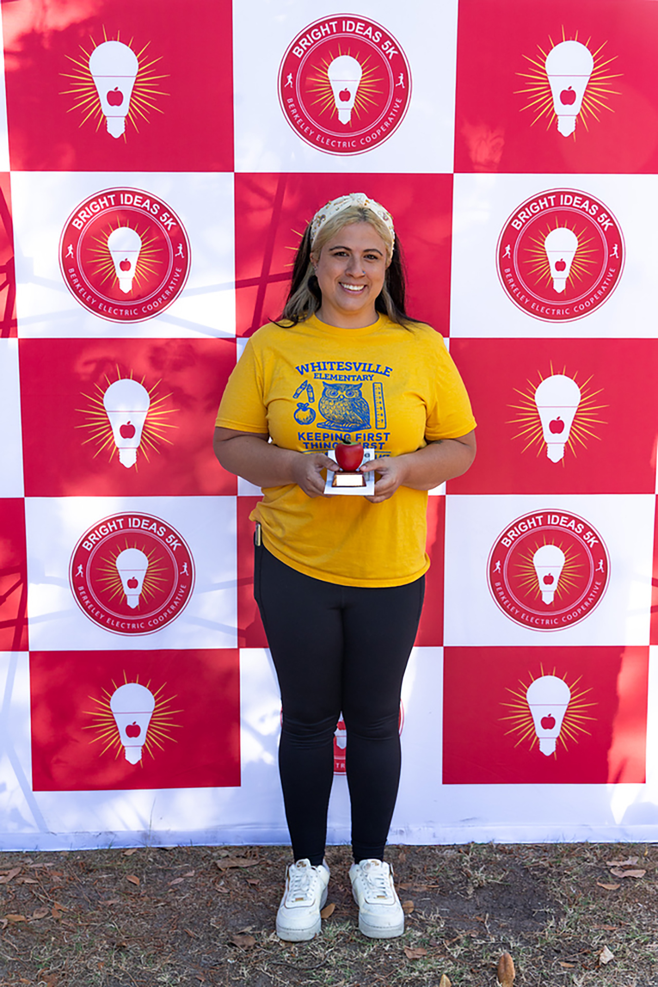 A woman holds a trophy in front of a red and white checkered backdrop with yellow light bulbs.