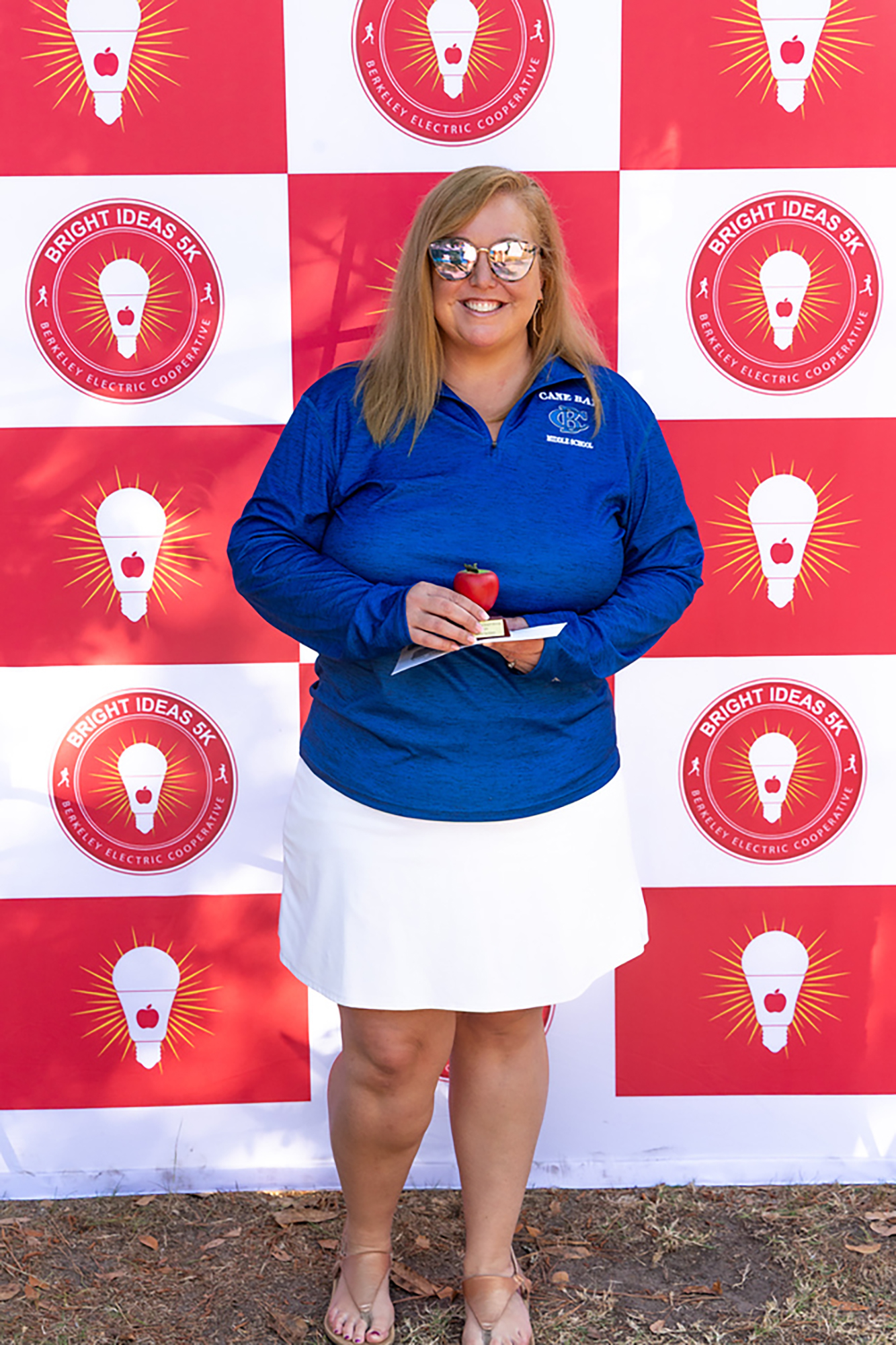 Woman in a blue long-sleeve shirt and white skirt holding an apple, standing in front of a red and white wall with logos.