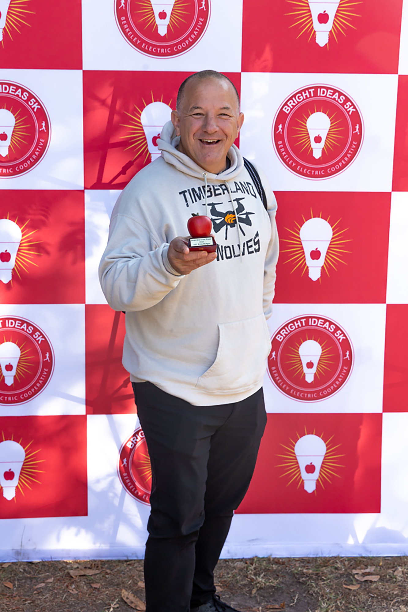 A person in a white hoodie smiles, holding a red apple, in front of a red and white background with lightbulbs.