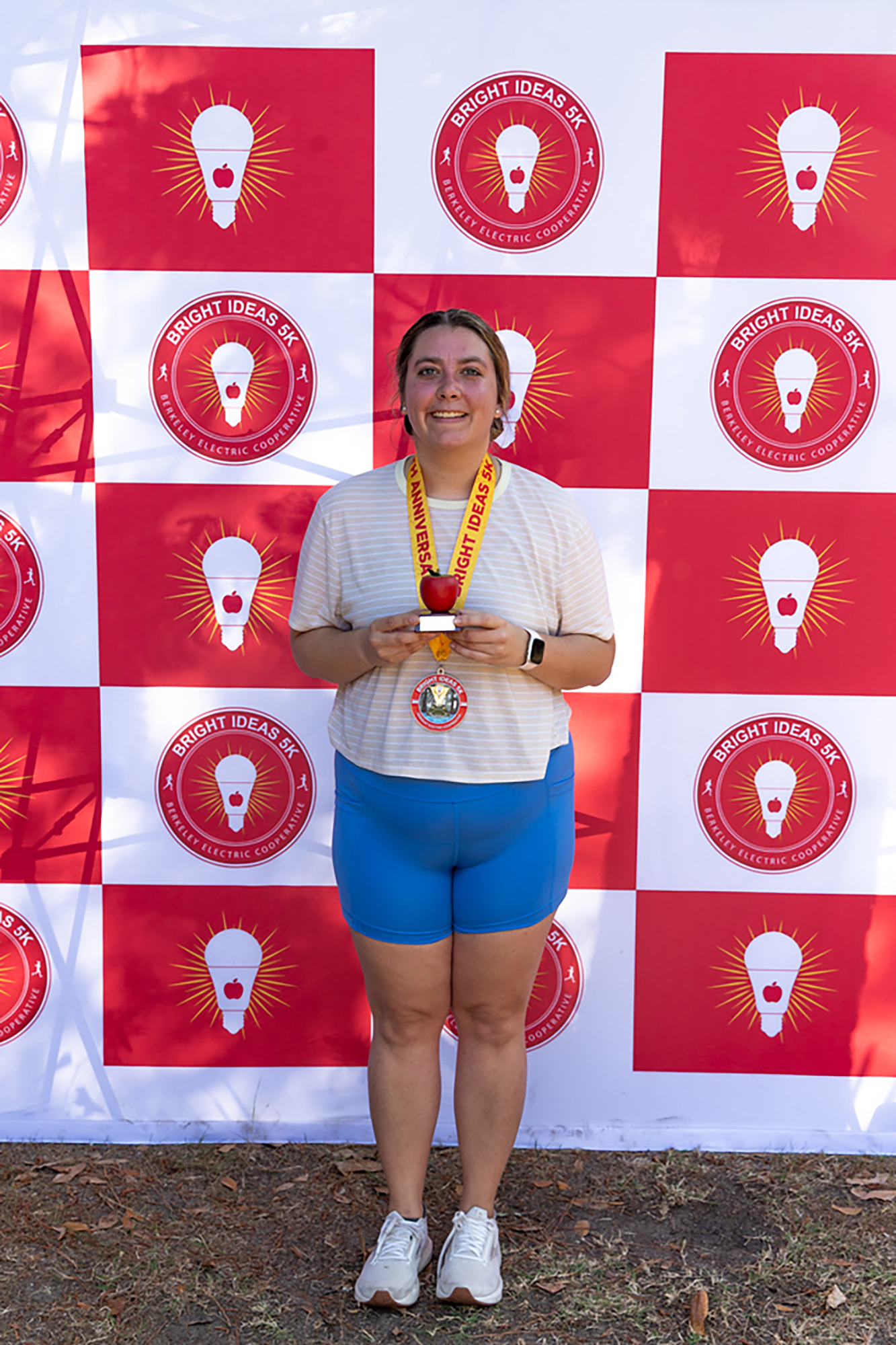 Woman in blue shorts and white sneakers stands before a red and white checkered wall with light bulb logos, holding a medal.