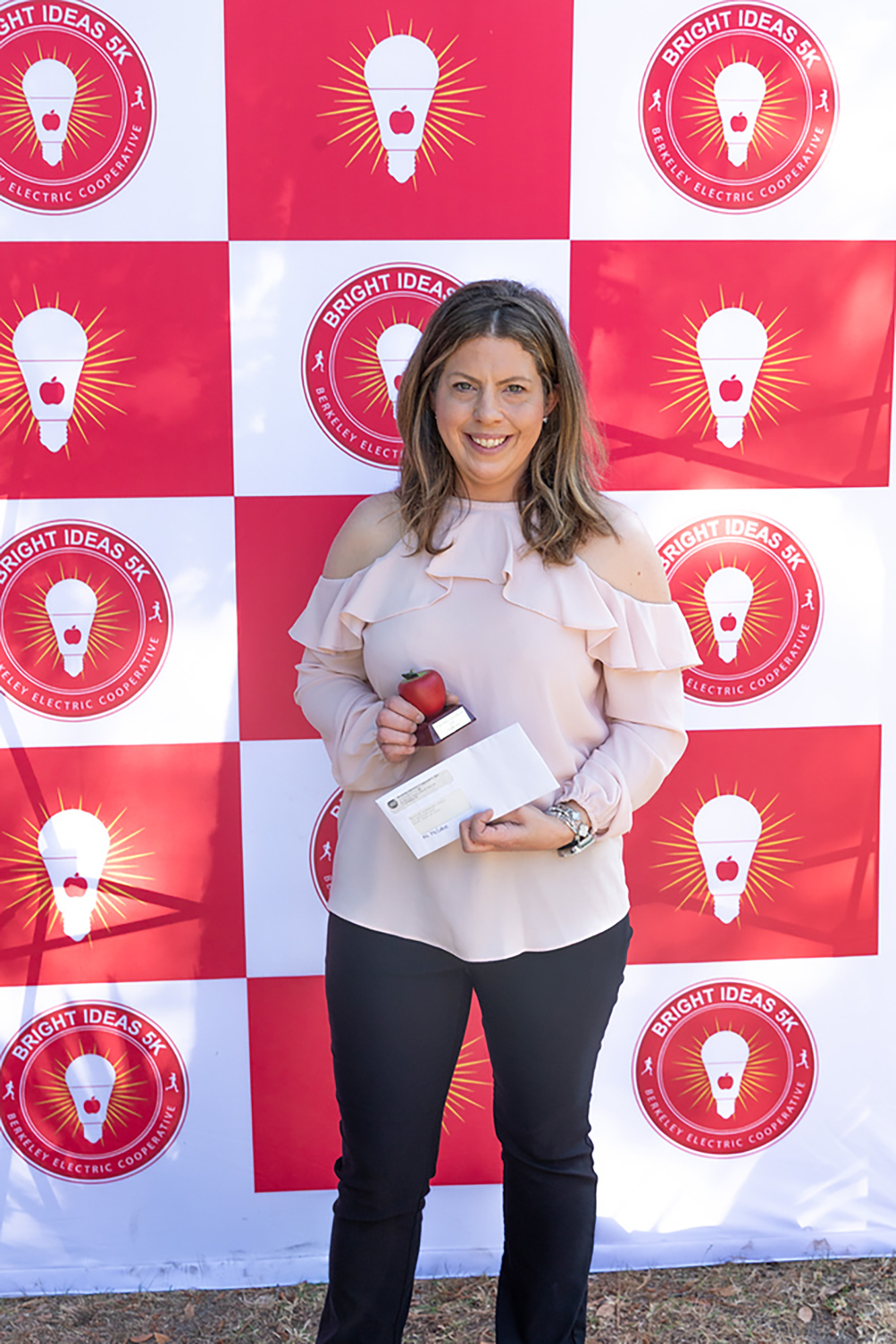 Woman stands before a wall with red and white squares, each with a lightbulb logo. She holds a small red trophy and a certificate.