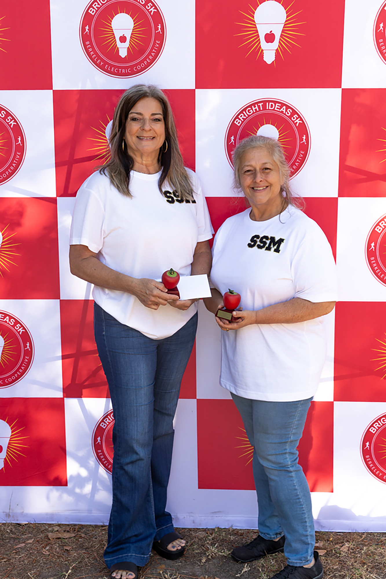 Two women stand before a red and white checkered backdrop with light bulbs. Each holds a small red object.