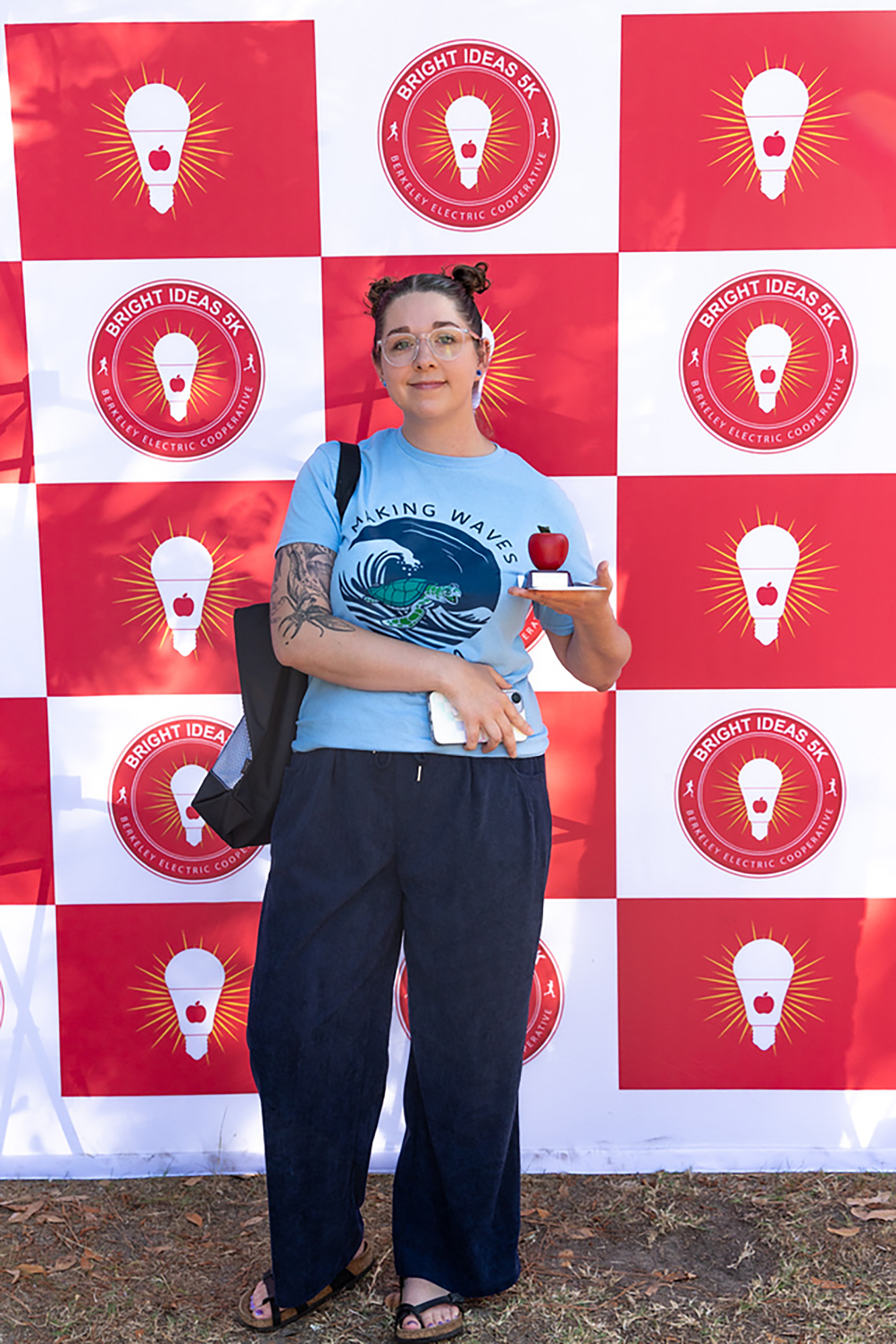 A person in a light blue shirt, holding an apple, stands in front of a wall with a repeating red and white pattern.