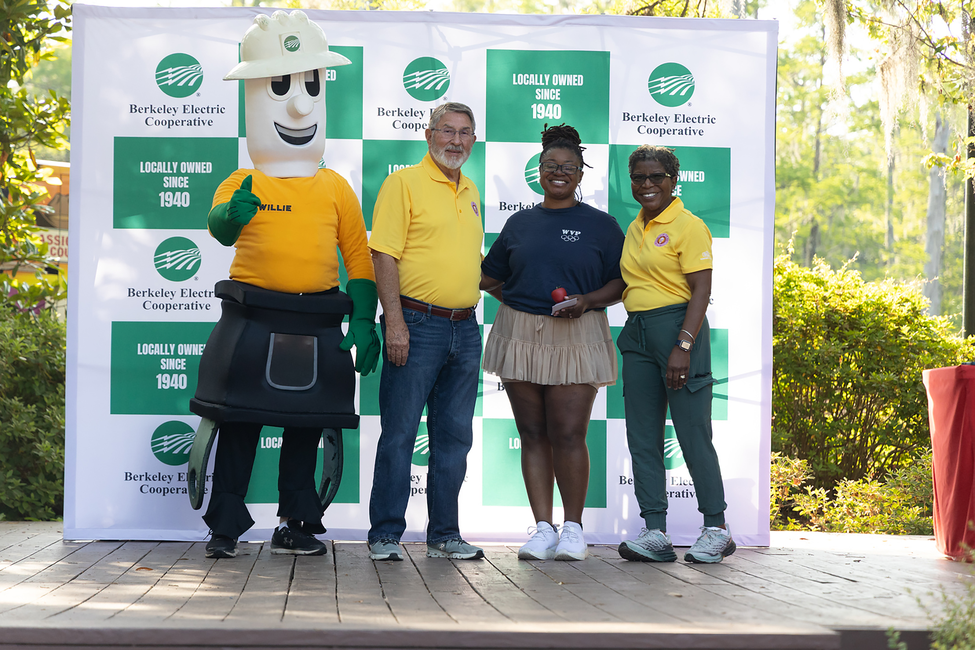 Four people on a stage. One person is in a costume. The others wear yellow shirts. A sign is behind them.