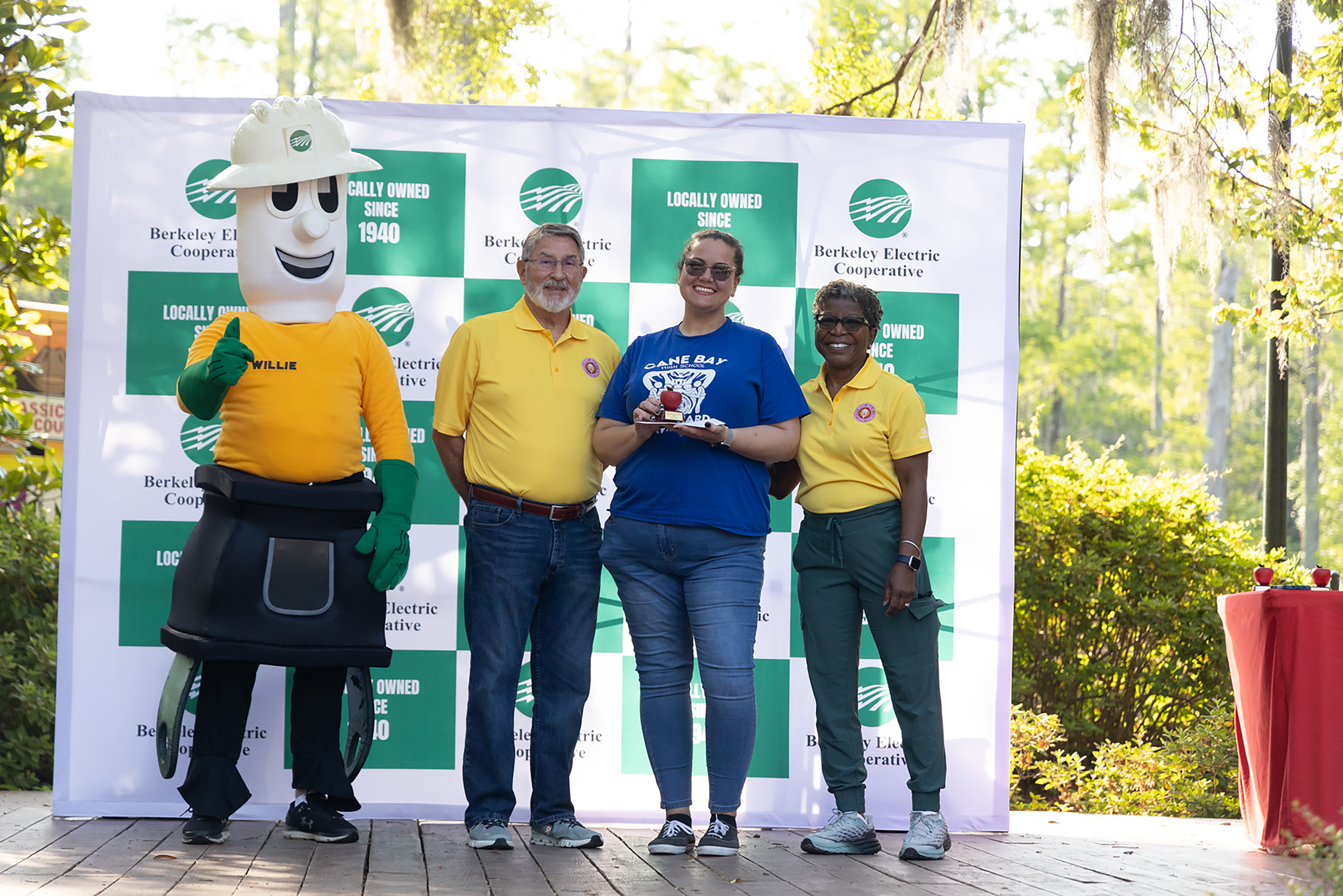 Four people, including a mascot, stand on a stage, with a backdrop of green and white signs.
