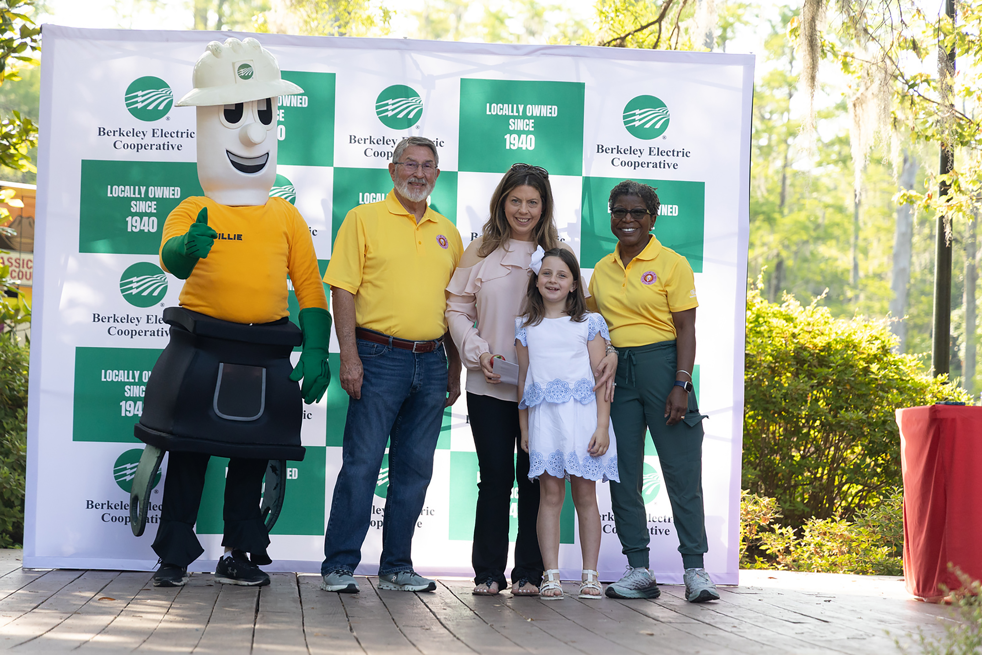 A group of people, including two adults, a child, and a mascot, stand together in front of a banner.