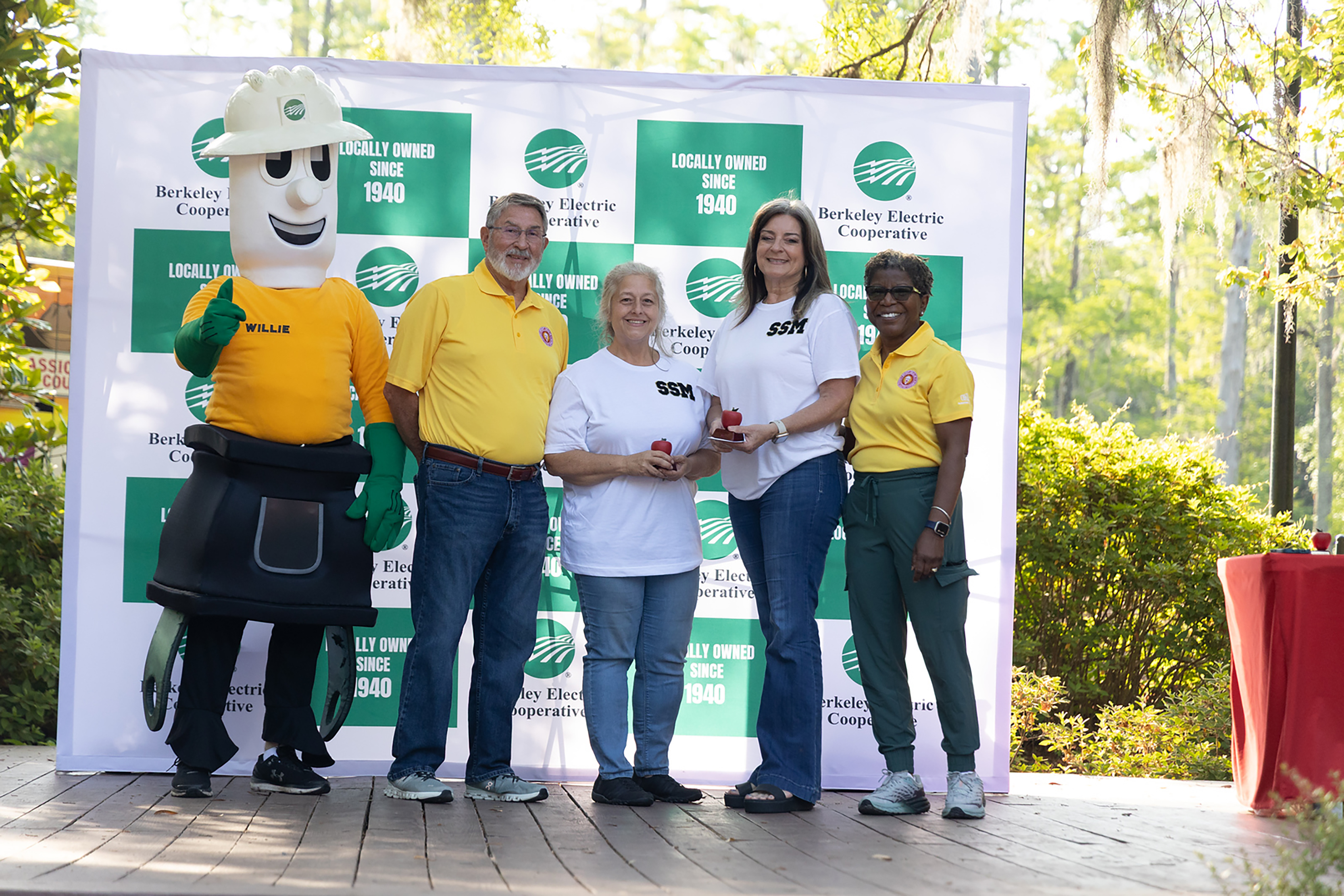 Five people stand on a wooden platform in front of a banner. The mascot wears a helmet and a yellow shirt. The others wear yellow shirts and pants.