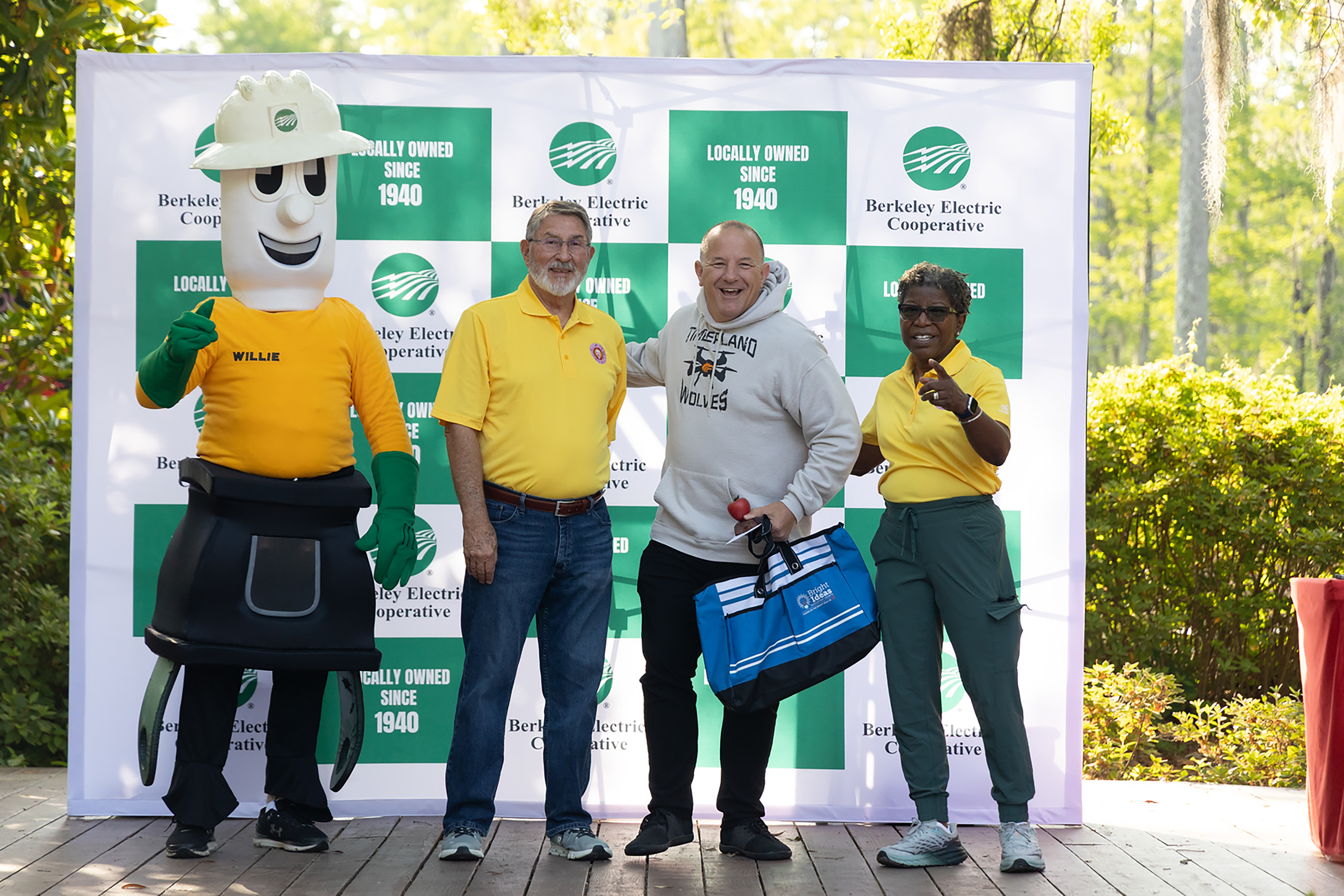 Four people in yellow shirts stand in front of a backdrop with green and white panels. One holds a blue bag.