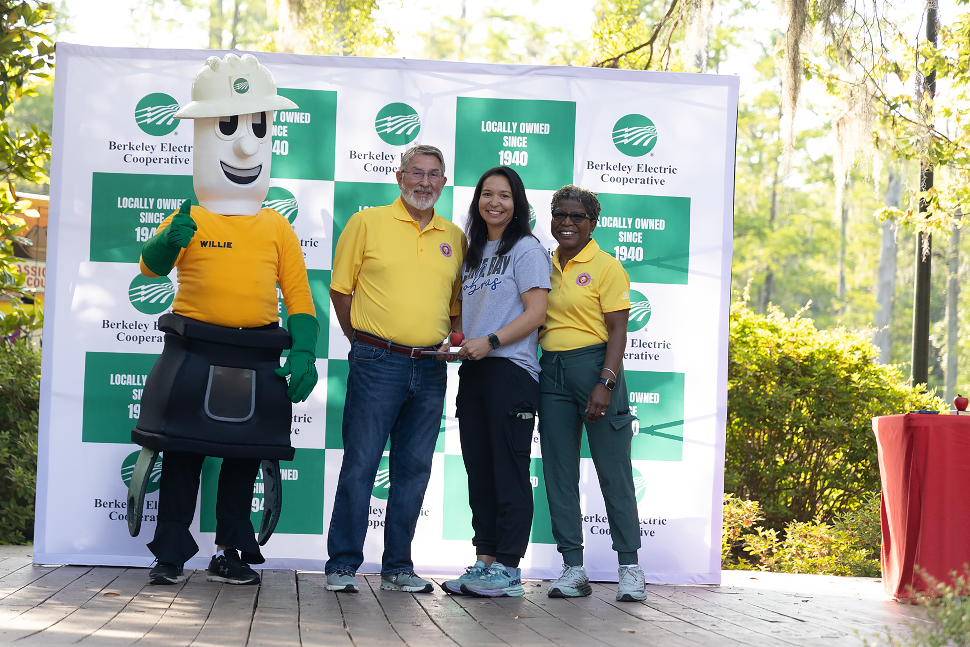 Four people in yellow shirts, one wearing a garden mascot costume, stand in front of a green and white backdrop.
