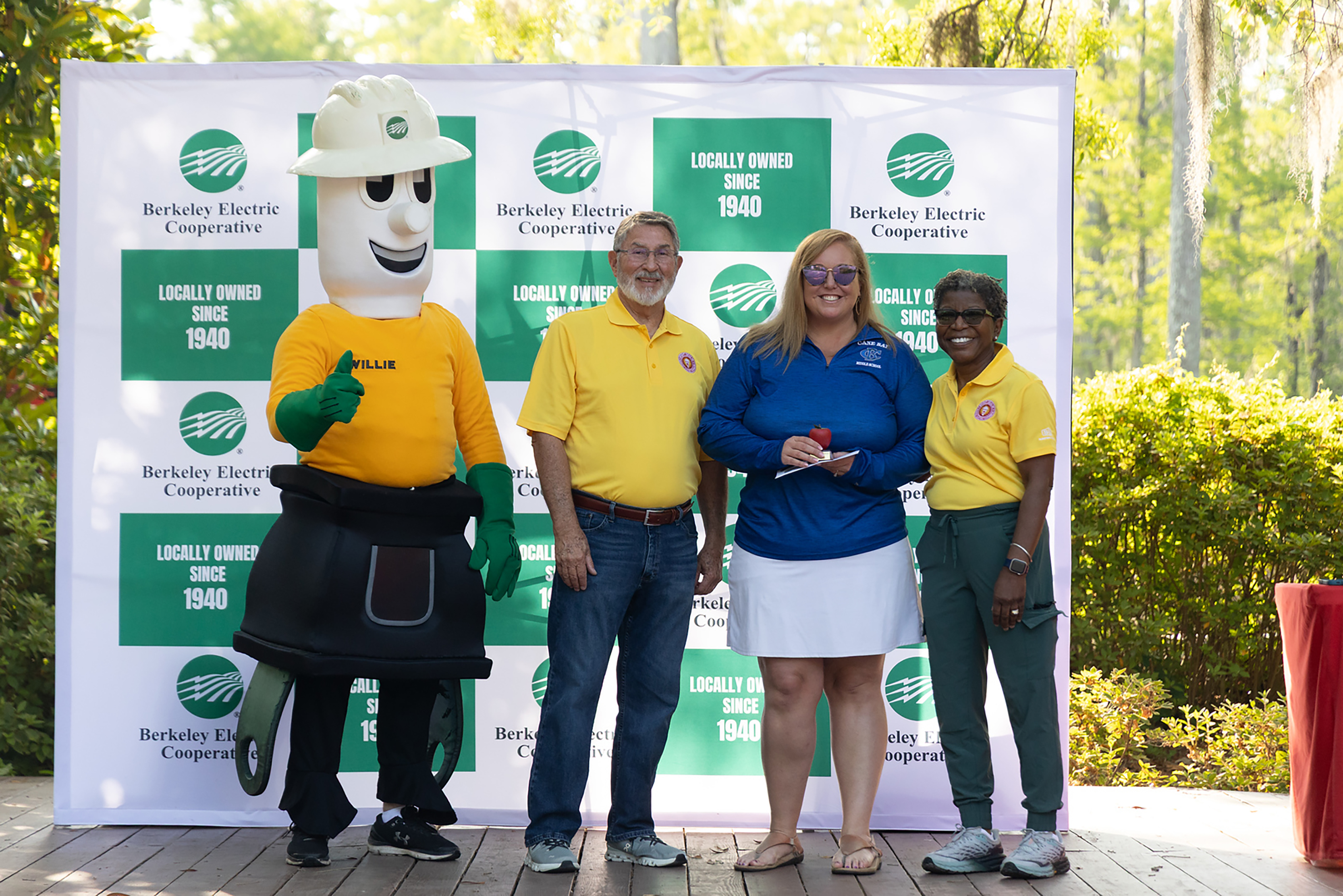 Four people in yellow shirts and one in a green costume stand on a stage. A green and white backdrop is behind them.