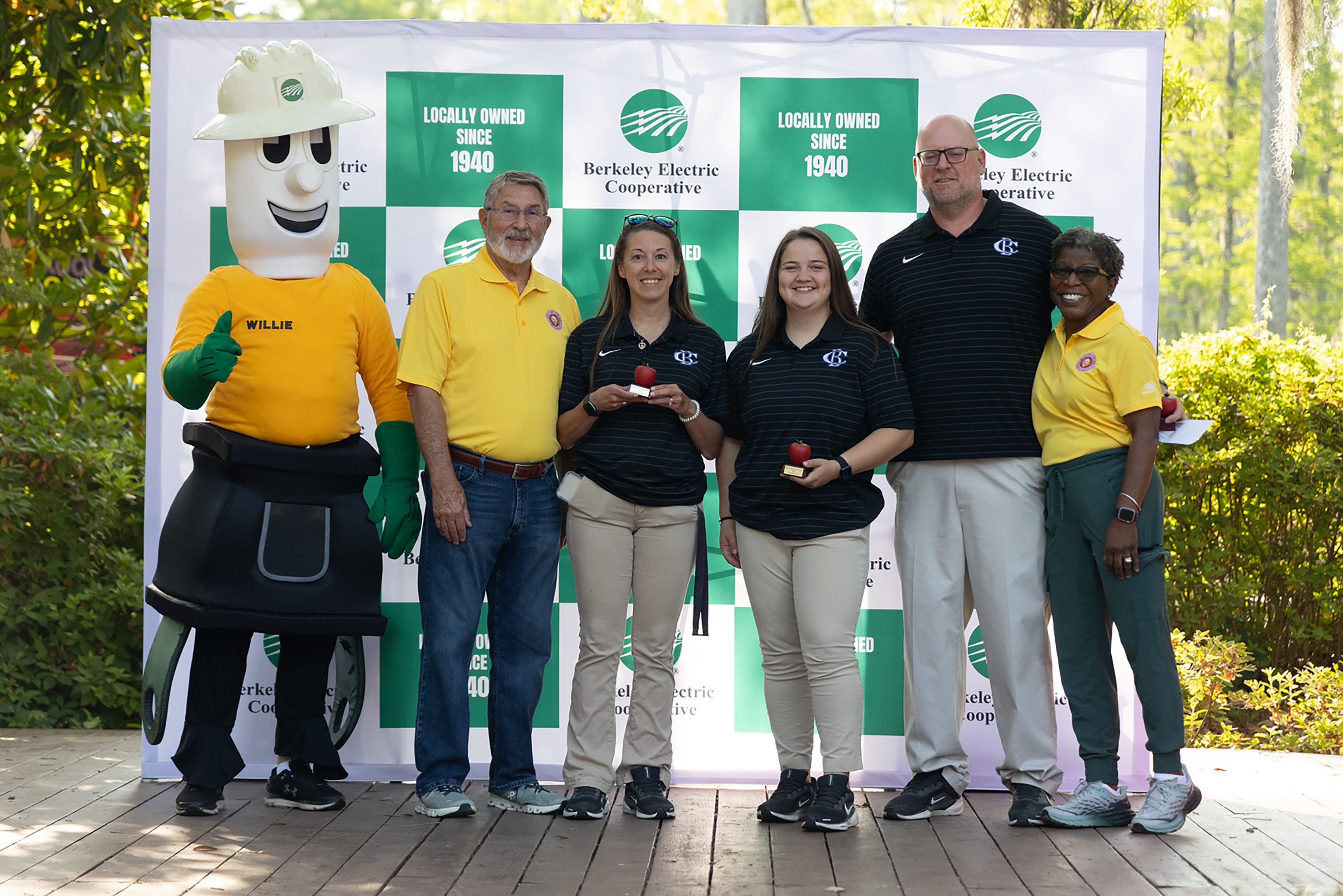 Five individuals, including a person in a mascot costume, stand together in front of a green and white backdrop.