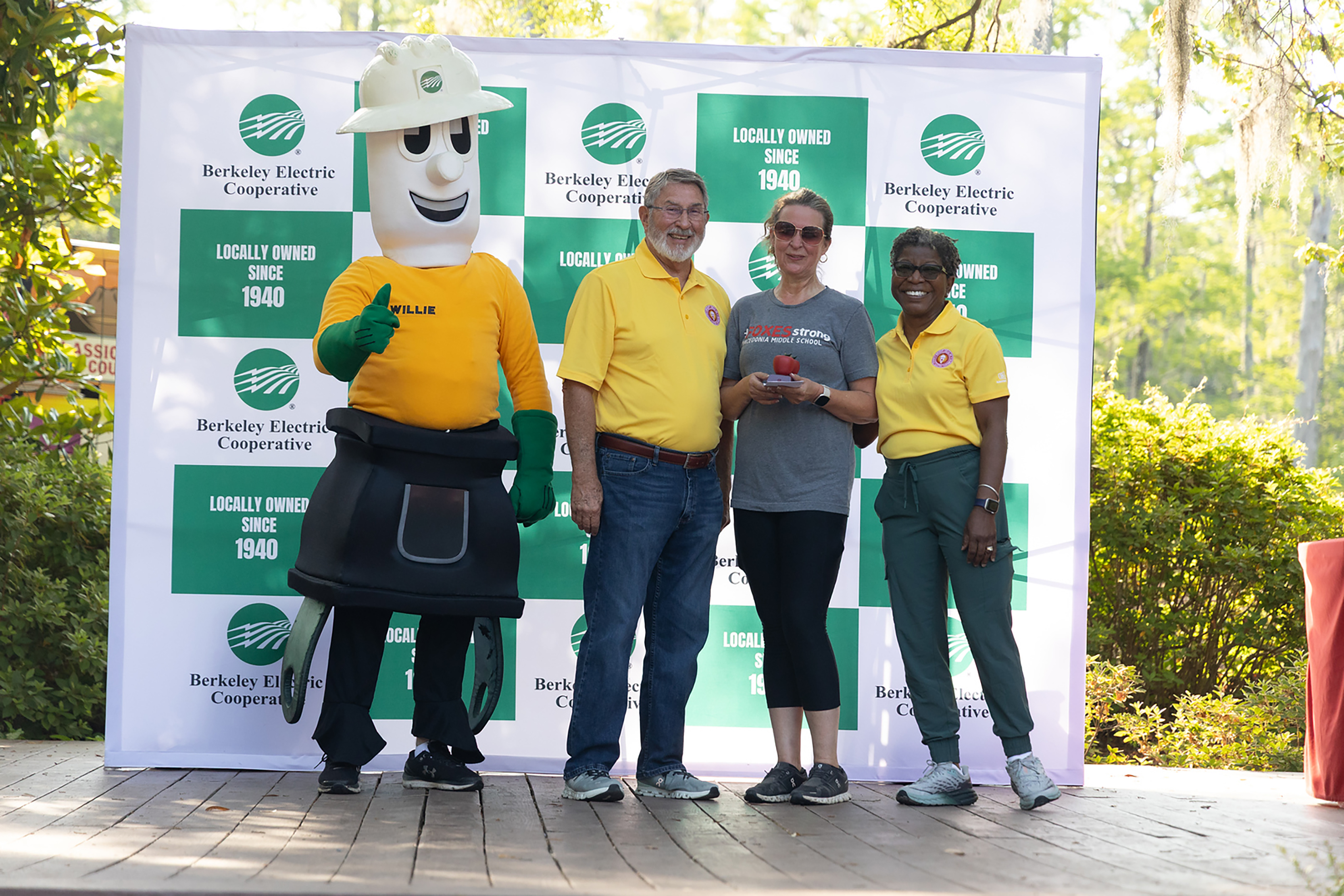 Five people stand in front of a large green-and-white sign with a mascot. Two women and one man wear yellow shirts.