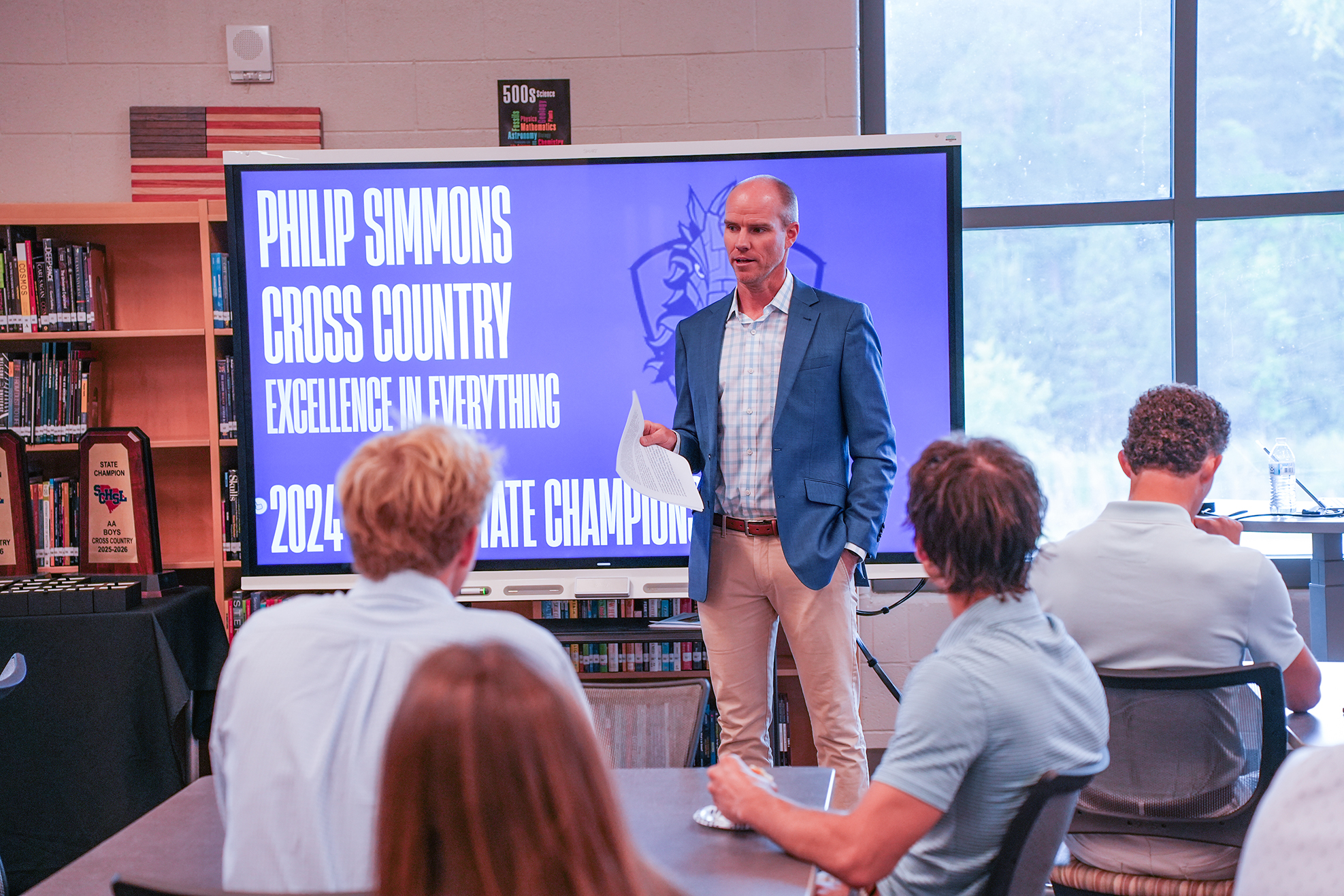 A man stands in a classroom, speaking to students seated around a table. A screen behind him displays the words "Philip Simmons Cross Country Excellence in Everything."