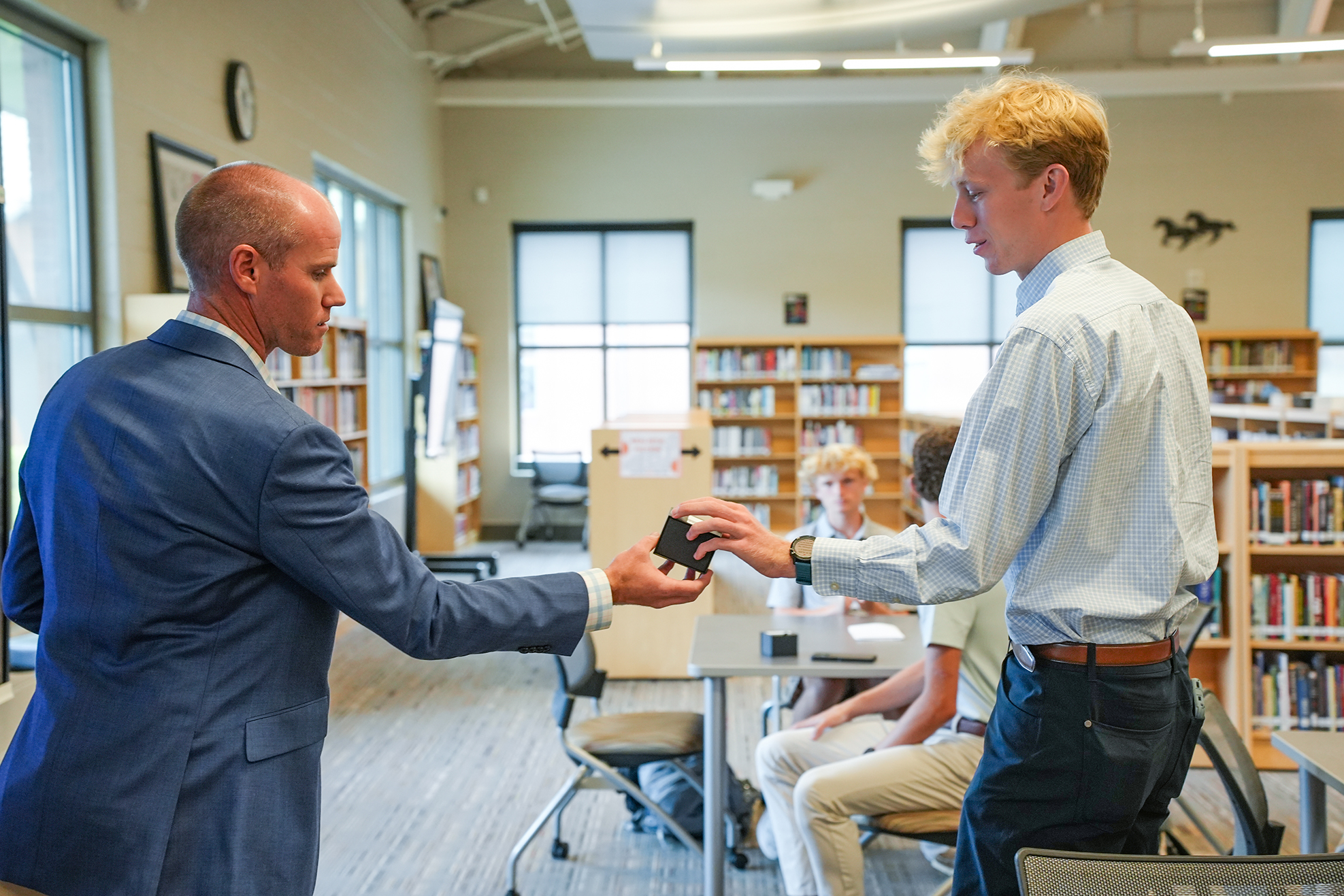 Two men in business attire exchange a small electronic device in a room with bookshelves and tables.