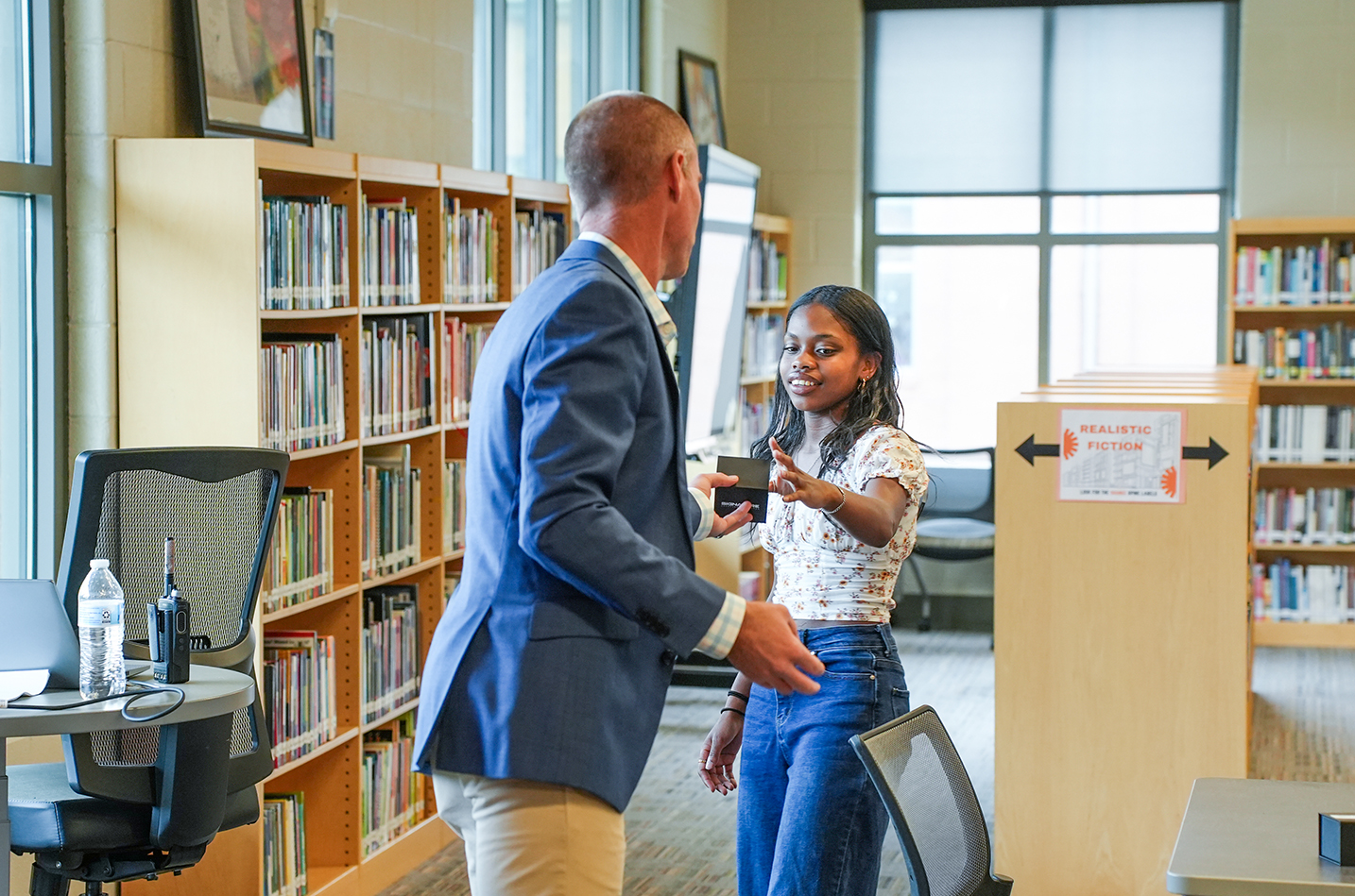 A man and a woman are standing in a library, conversing. The woman holds a tablet, and a bookshelf is behind them.