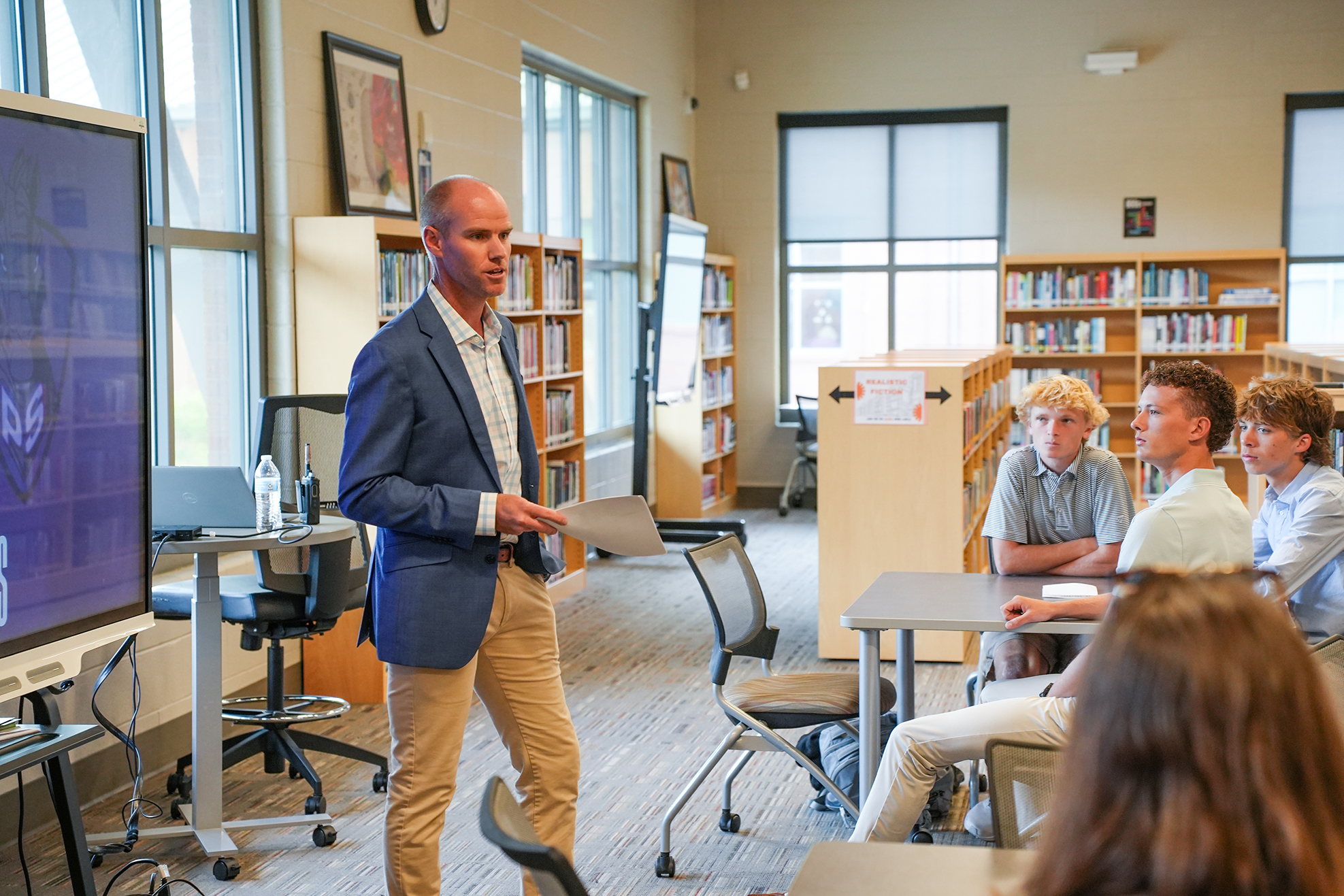 Man stands in library, speaking to seated students around a table with a large screen displaying a figure.