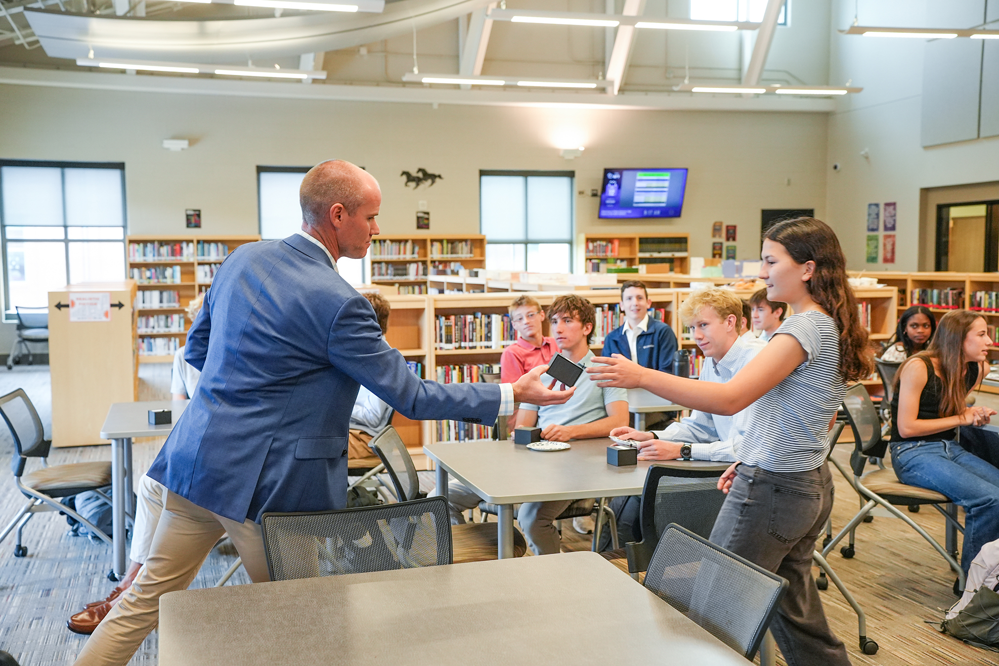A man and woman interact in a library with tables, chairs, and people seated or standing.