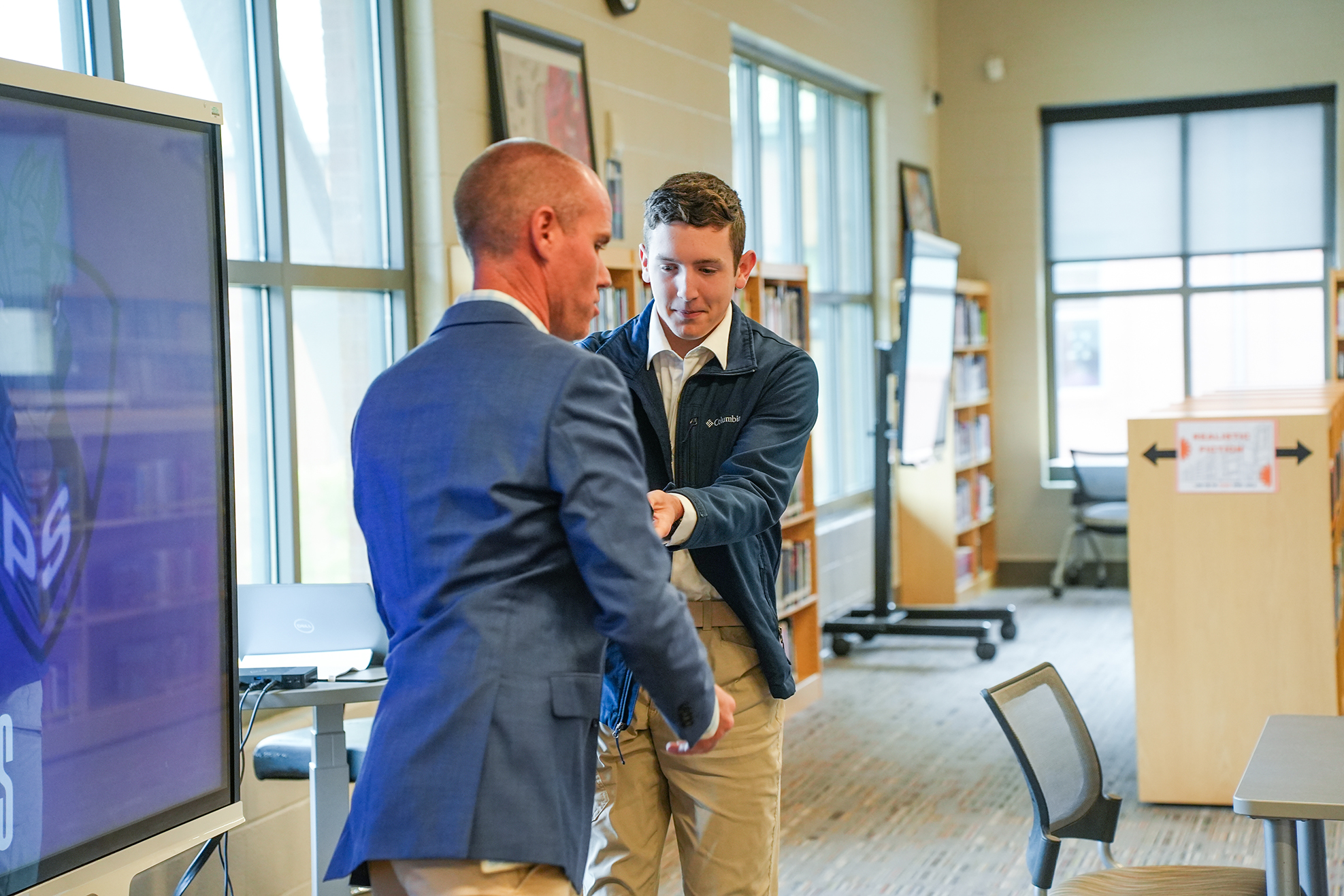 Two men in suits inside a classroom with a monitor, shelves, and chairs. Sunlight enters through large windows.