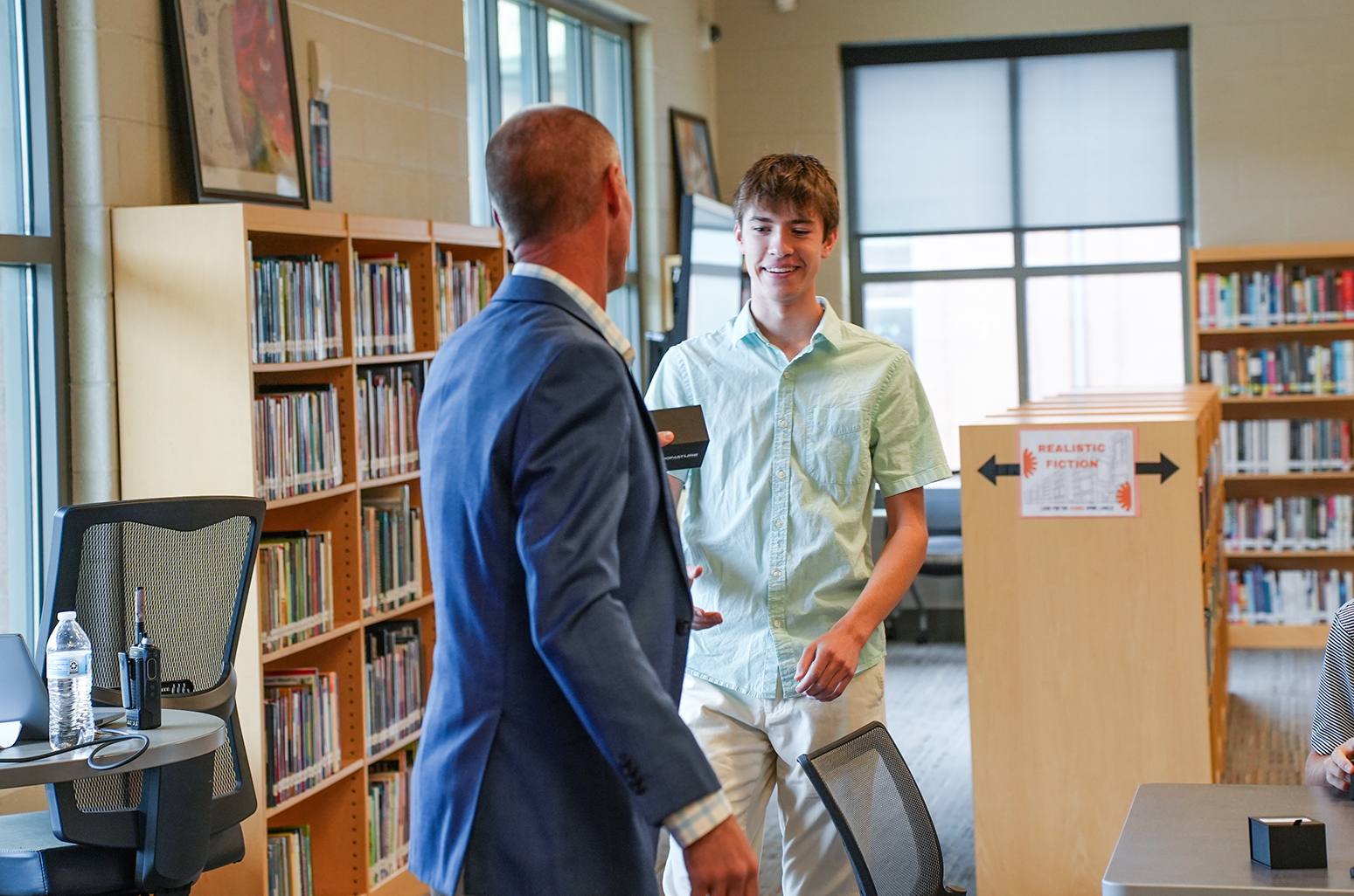 Two people, one in a light blue suit, the other in a light green shirt, talk in a library.