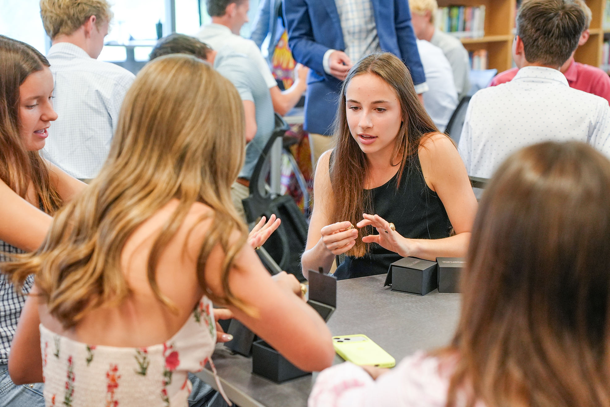 A group of students sitting at a table, engaging in conversation. Some students wear casual clothing.