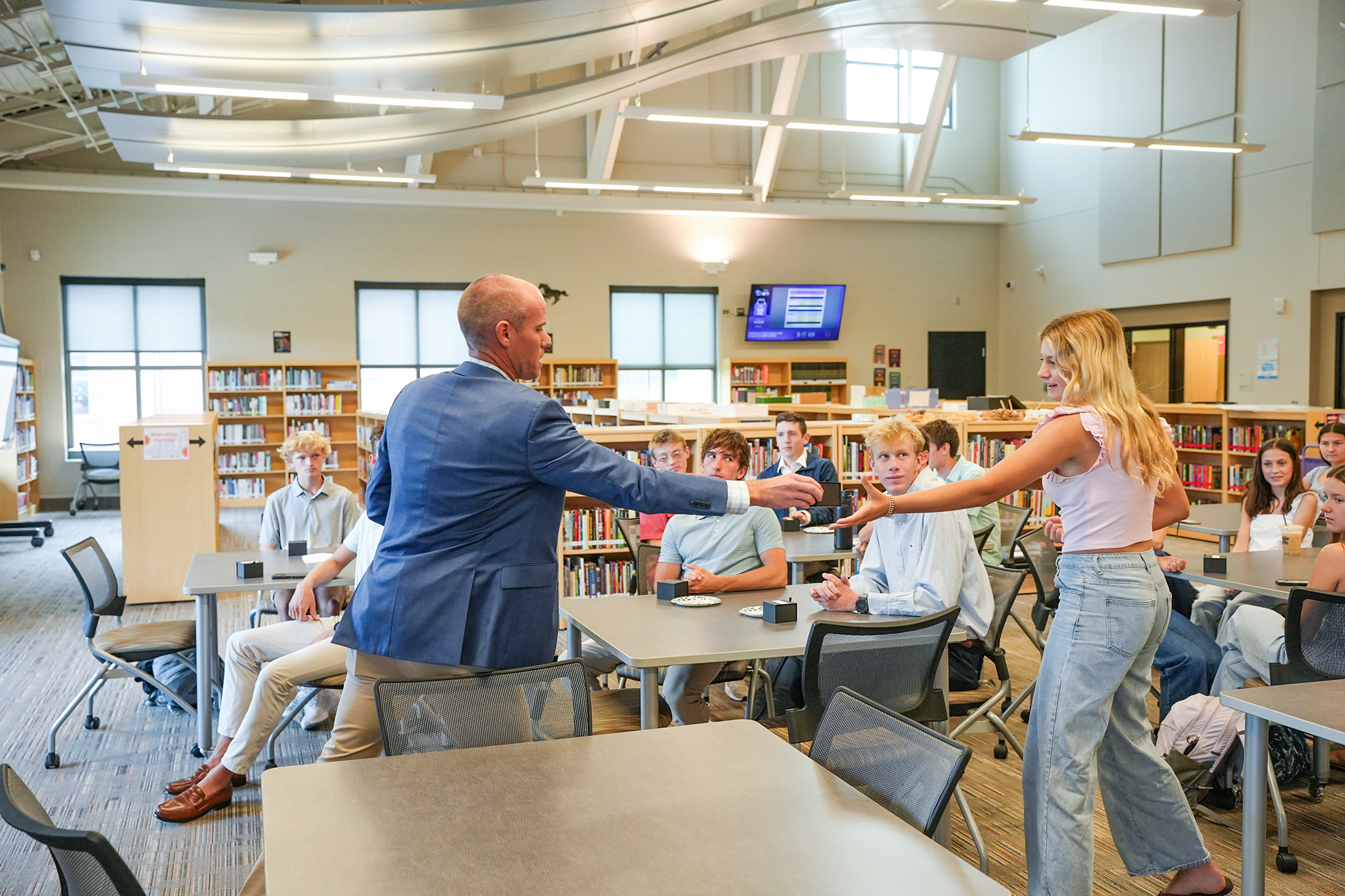 Two people in a classroom setting, with the man in blue shaking the woman's hand. Behind them, people sit at tables.