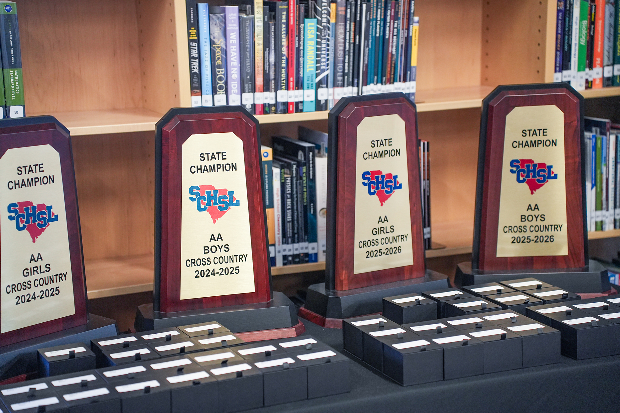 Trophies for Boys' and Girls' Cross Country arranged on a table with a bookshelf filled with books in the background.