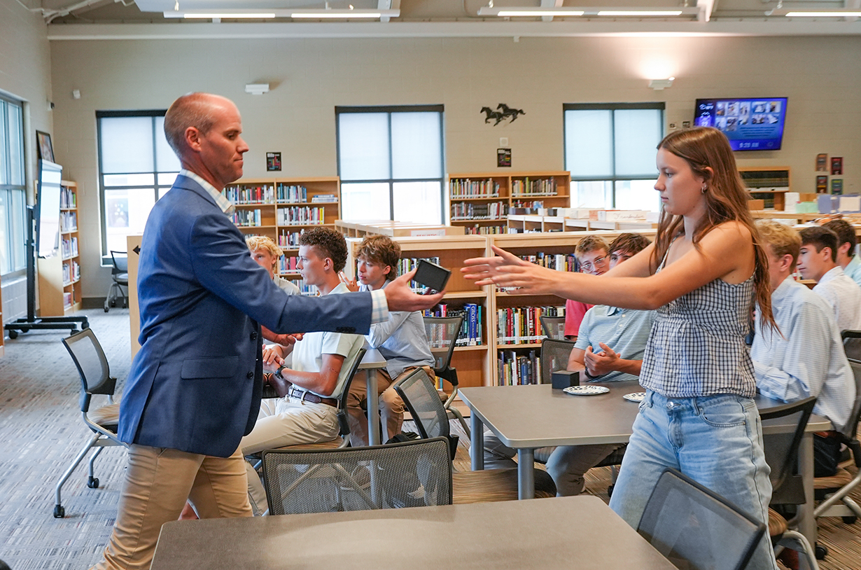 A man and woman are facing each other while gesturing in a classroom with bookshelves. Students sit at tables.