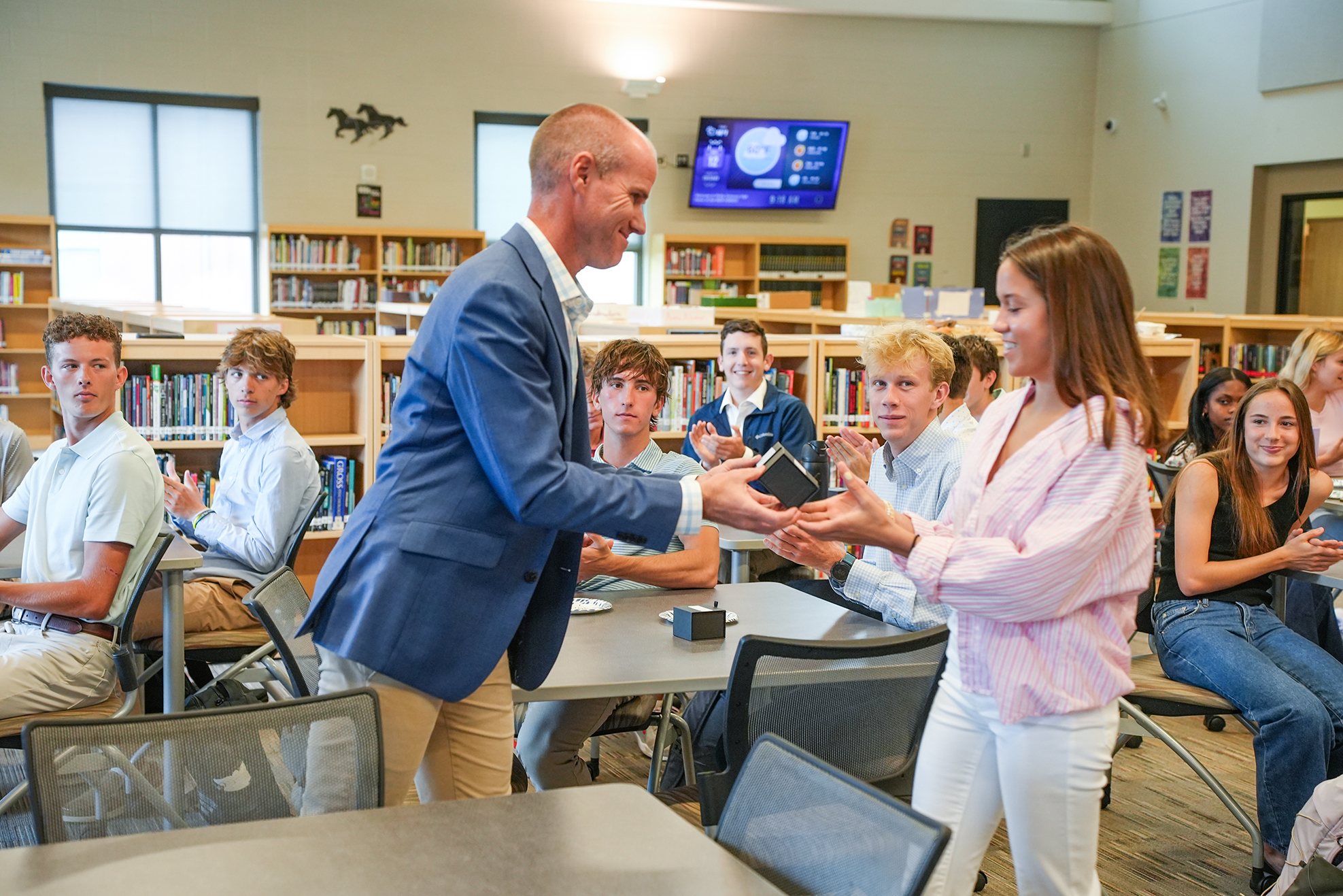 A man in a blue suit hands an award to a woman in a pink top in a classroom.