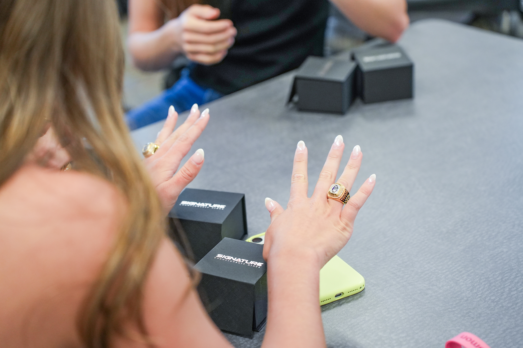 A person with hands raised and long hair at a table with a cellphone, boxes, and another person.