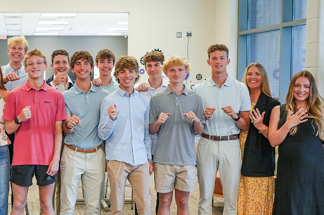 Group of young adults in light-colored shirts and shorts, some wearing belts, pose in an indoor room.