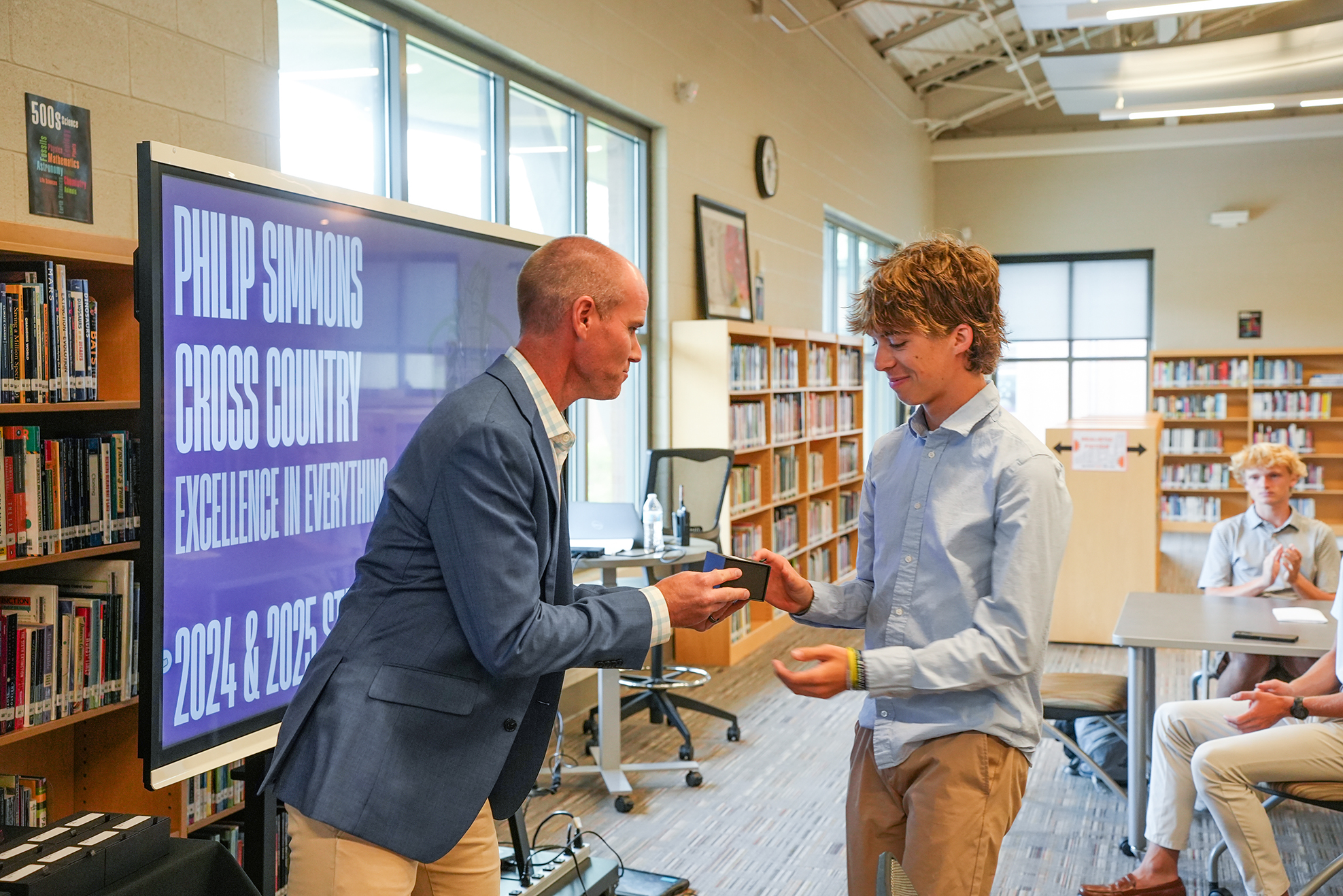 Two individuals in a library with bookshelves; one in a suit hands a tablet to a student.