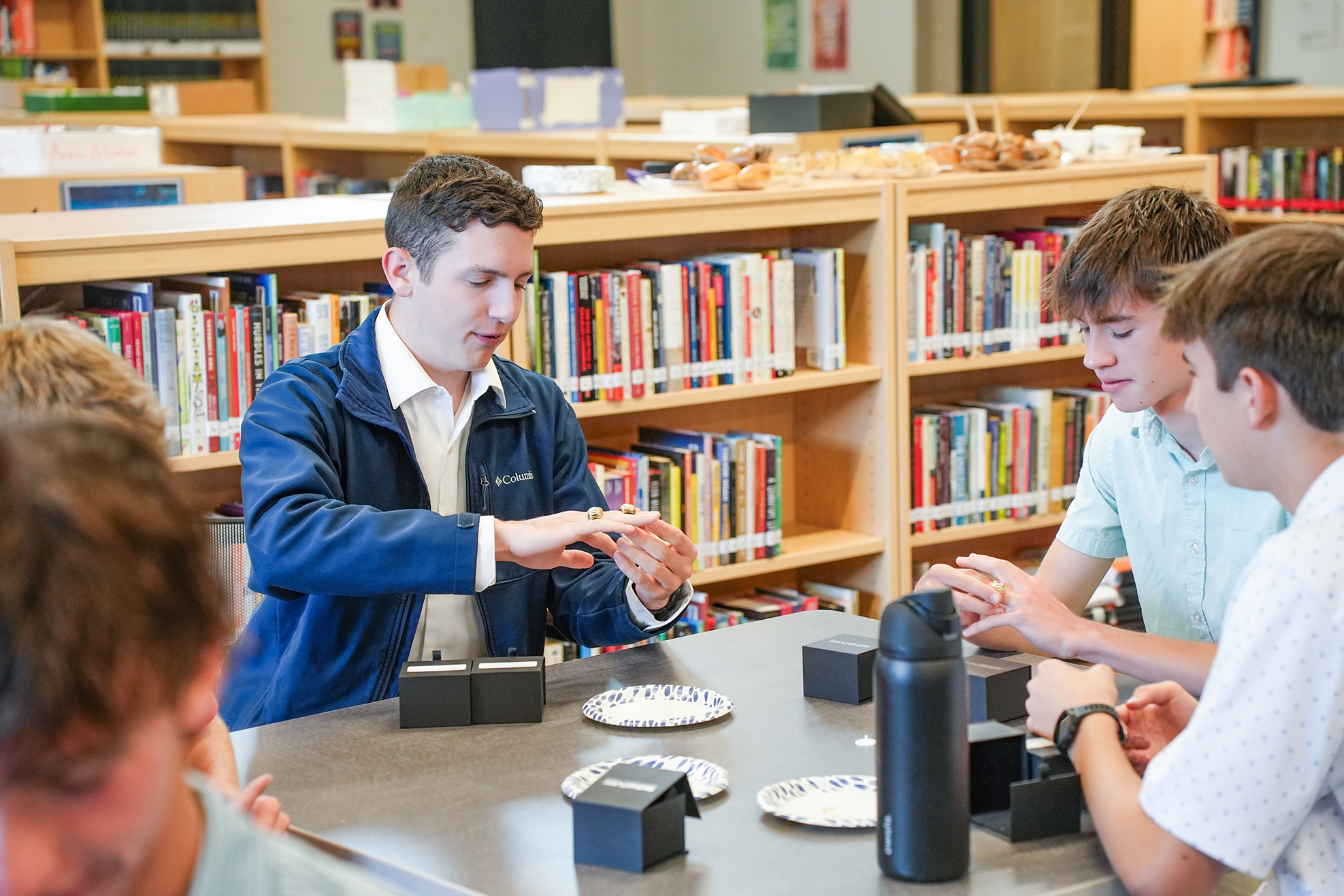 Five students sit at a table in a library, with one person speaking. Bookshelves with books are in the background.