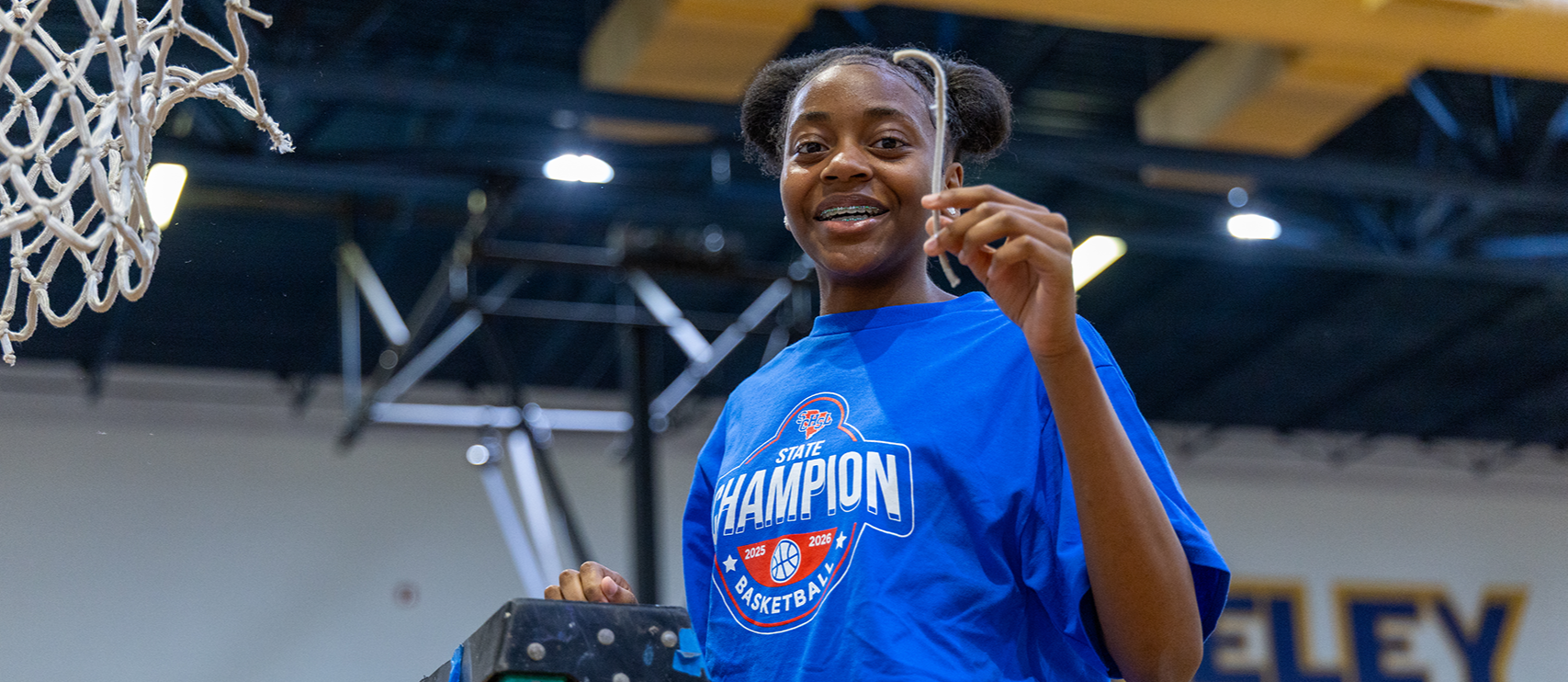 A woman in a blue shirt stands on a ladder, smiling, with a basketball hoop and a "Champion" logo in the background.