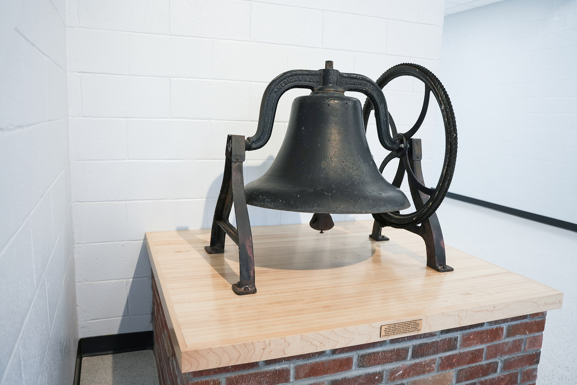 A black bell with a crank on a wooden platform atop a red brick pillar. A white wall is behind the platform.
