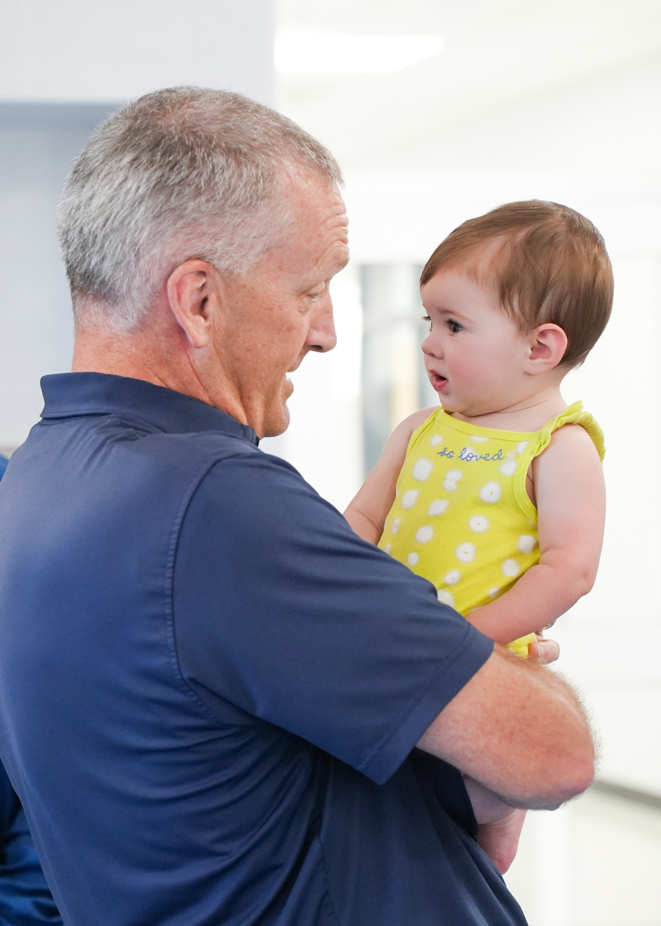 An older man with gray hair holds a baby girl with short red hair. The baby wears a yellow sleeveless top.