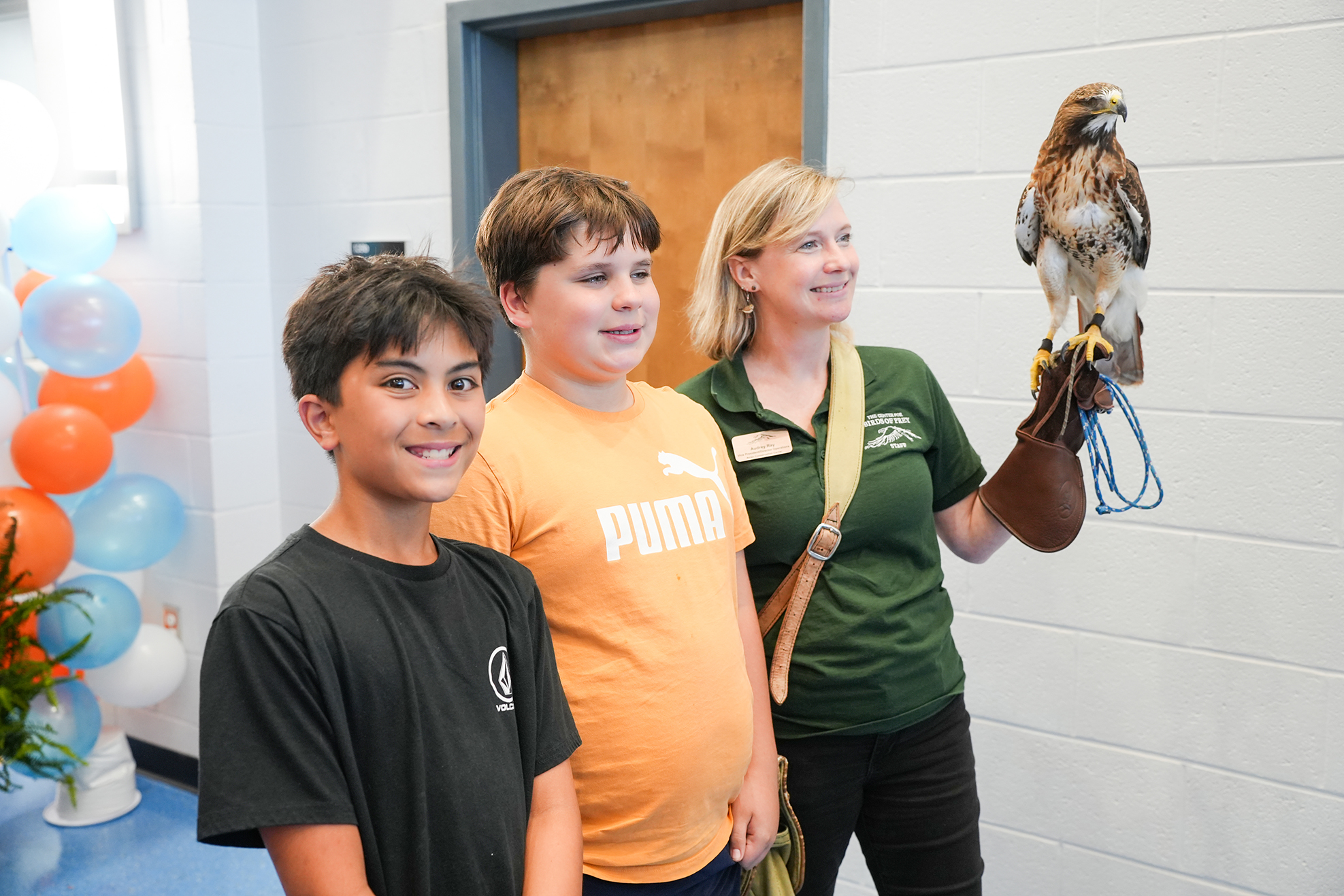 Three people standing in a room. A woman holds a bird. Two boys in front, one with an orange shirt.