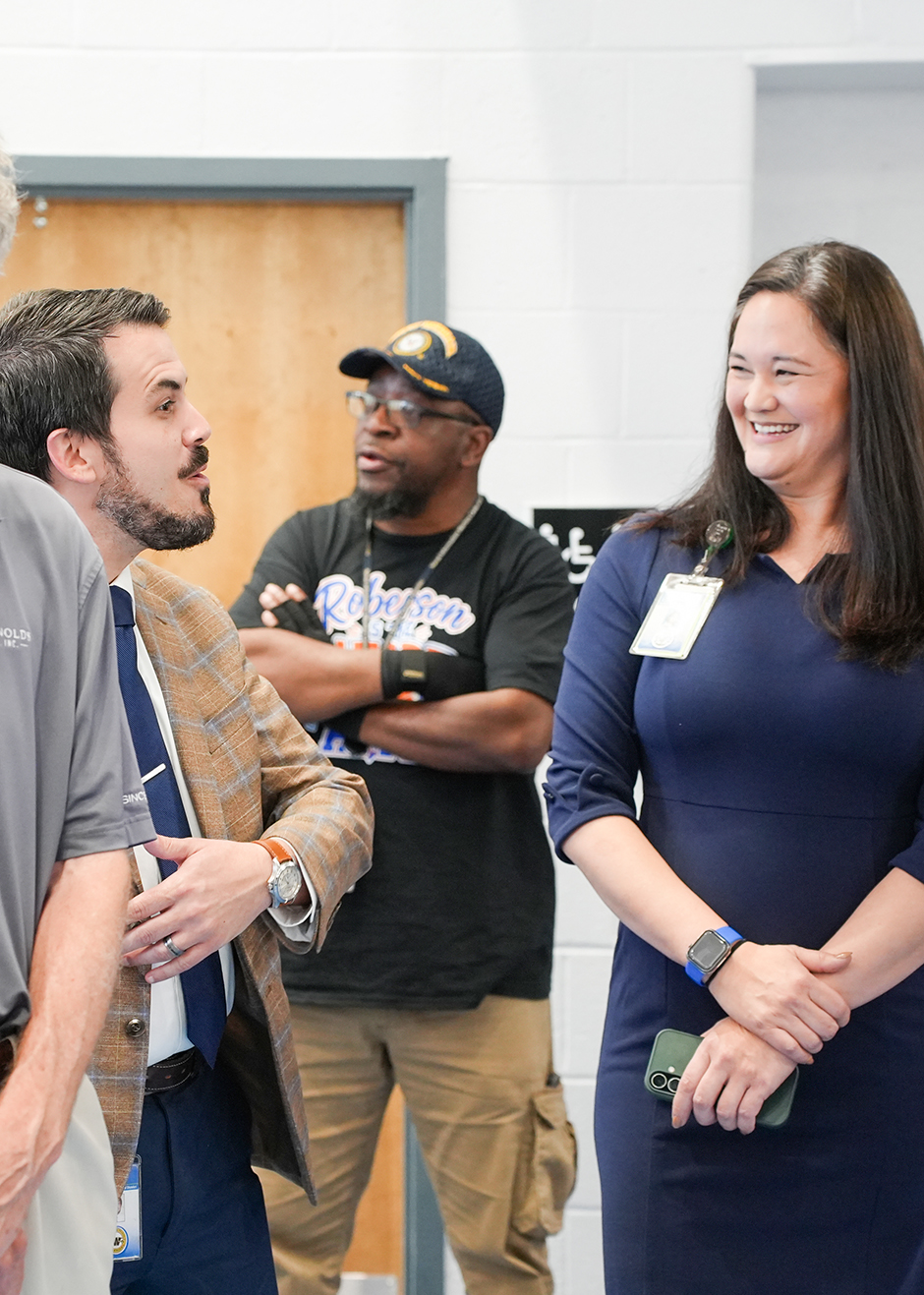 Four people stand in a room, with a woman in a blue dress smiling. Behind her, a man in a black shirt has crossed arms.