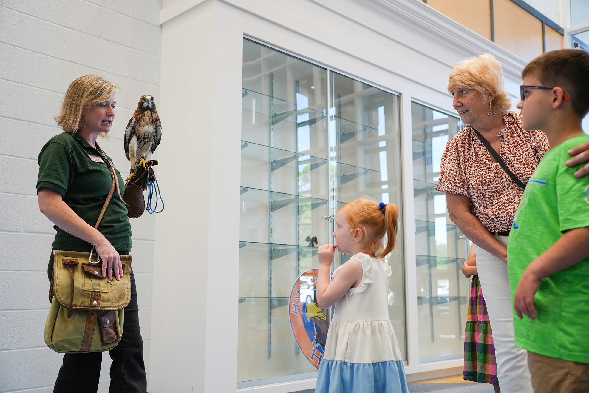 An adult woman with a hawk on her arm talks to a girl and boy in a white room.